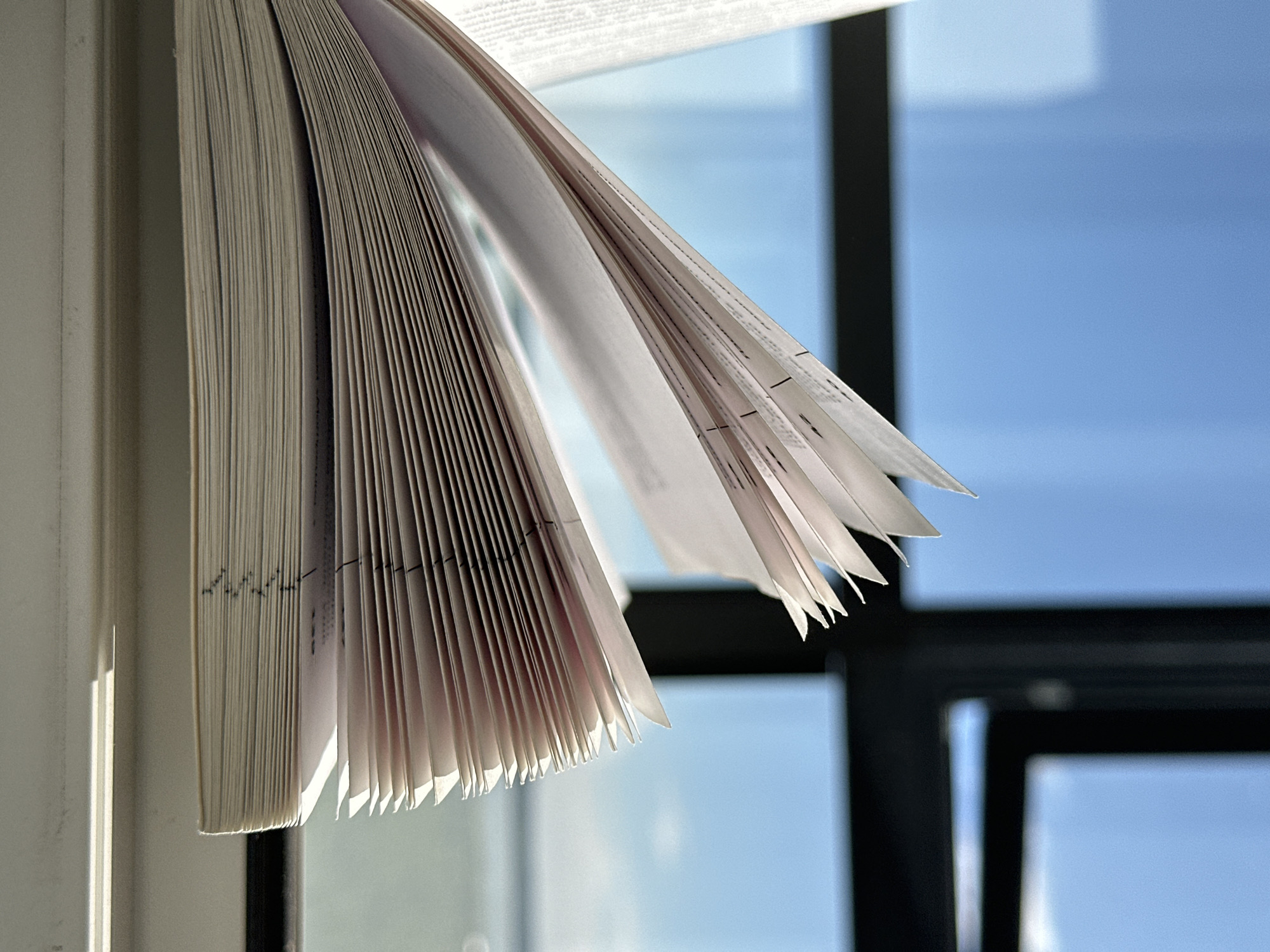 The image shows the edge of a thick book with its pages fanned out. The book is positioned near a window. Outside the window, there is a view of a blue sky. The window frame is black. The light and shadows suggest the photo was taken on a bright day. The location is Unknown, Russia. There are no people present in the image.