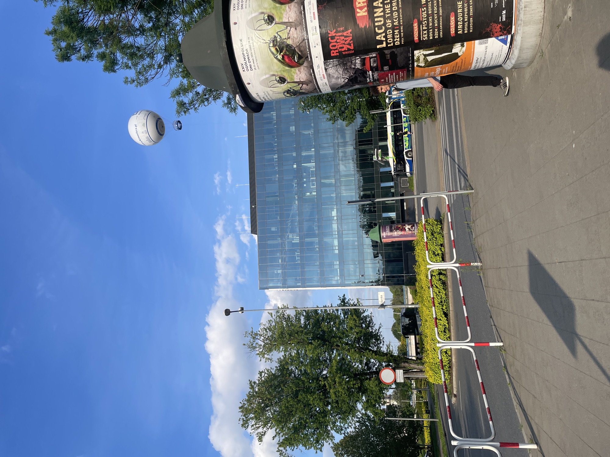 Here is a description of the image:
The image shows a street scene, likely in Poland, given the Polish text on the advertisements and the company Atmosfera.pl. A tall, modern glass building is in the background, reflecting the blue sky and white clouds. In the foreground, there is a cylindrical advertising kiosk covered in posters for various events including music festivals, insect-themed events, and concerts. A person is walking away from the kiosk. Roadside barriers are set up next to the sidewalk. A large, white inflatable sphere floats in the sky, likely advertising something. The overall scene depicts an urban environment on a sunny day.