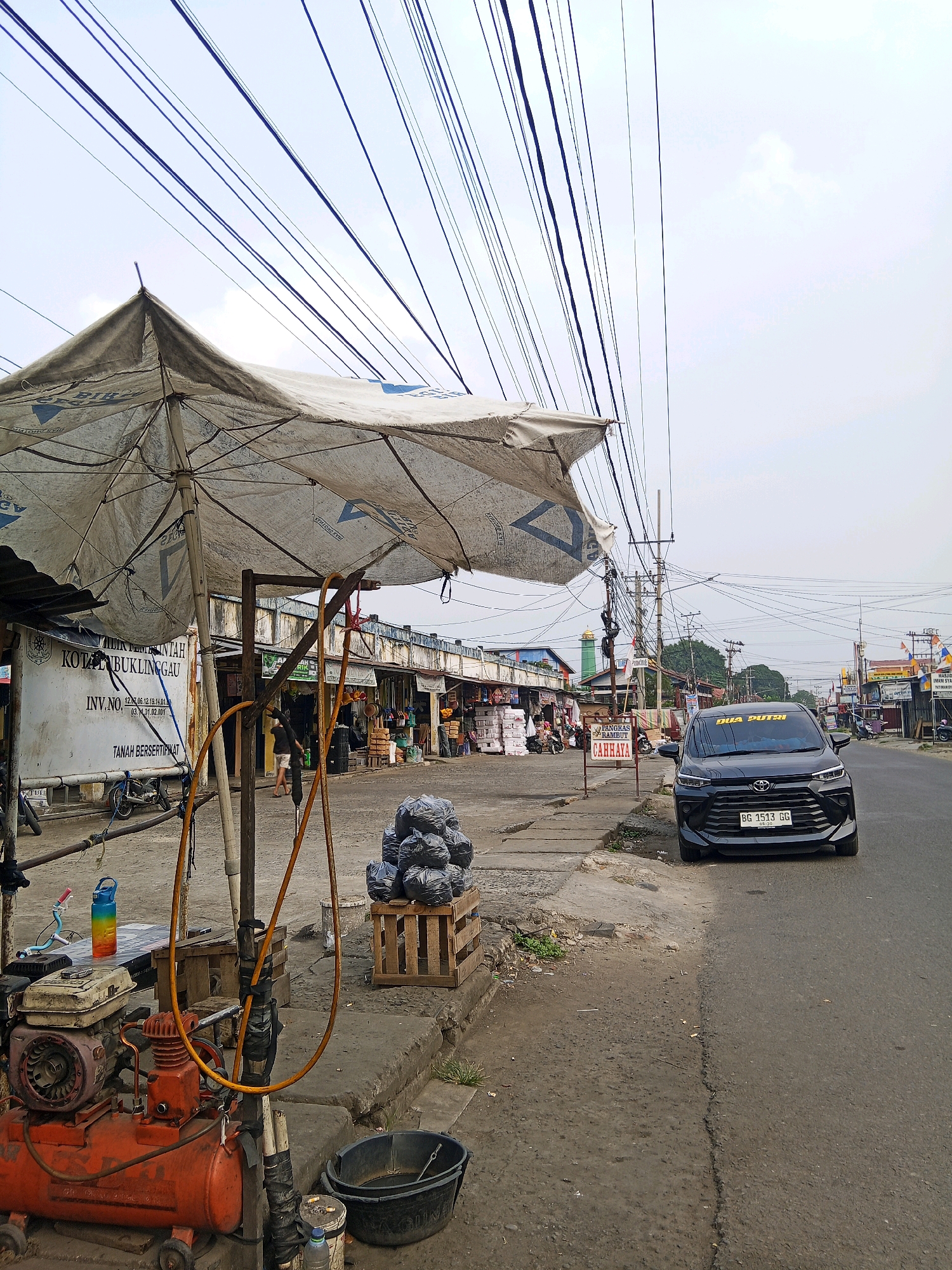 The image captures a street scene in Lubuklinggau, Indonesia, characterized by an overhead network of tangled electrical wires. In the foreground, a worn white tarp stretched over a metal frame, possibly a makeshift awning, shades a collection of objects. These include an orange air compressor with hoses, a colorful water bottle, and a black bucket. To the right, a stack of black trash bags sits on a wooden pallet.
The street itself appears to be paved, with a concrete sidewalk running alongside it. A black car with a license plate reading "BG 1513 GG" is parked on the right side of the road, facing forward. In the middle ground, several shops or stalls line the street, with various items displayed outside. People are visible in the distance, suggesting a bustling marketplace or commercial area. The architecture of the buildings appears to be simple, with storefronts facing the road. The overall atmosphere is that of an active, everyday street scene in a populated area. The sky is overcast, indicating a cloudy day.