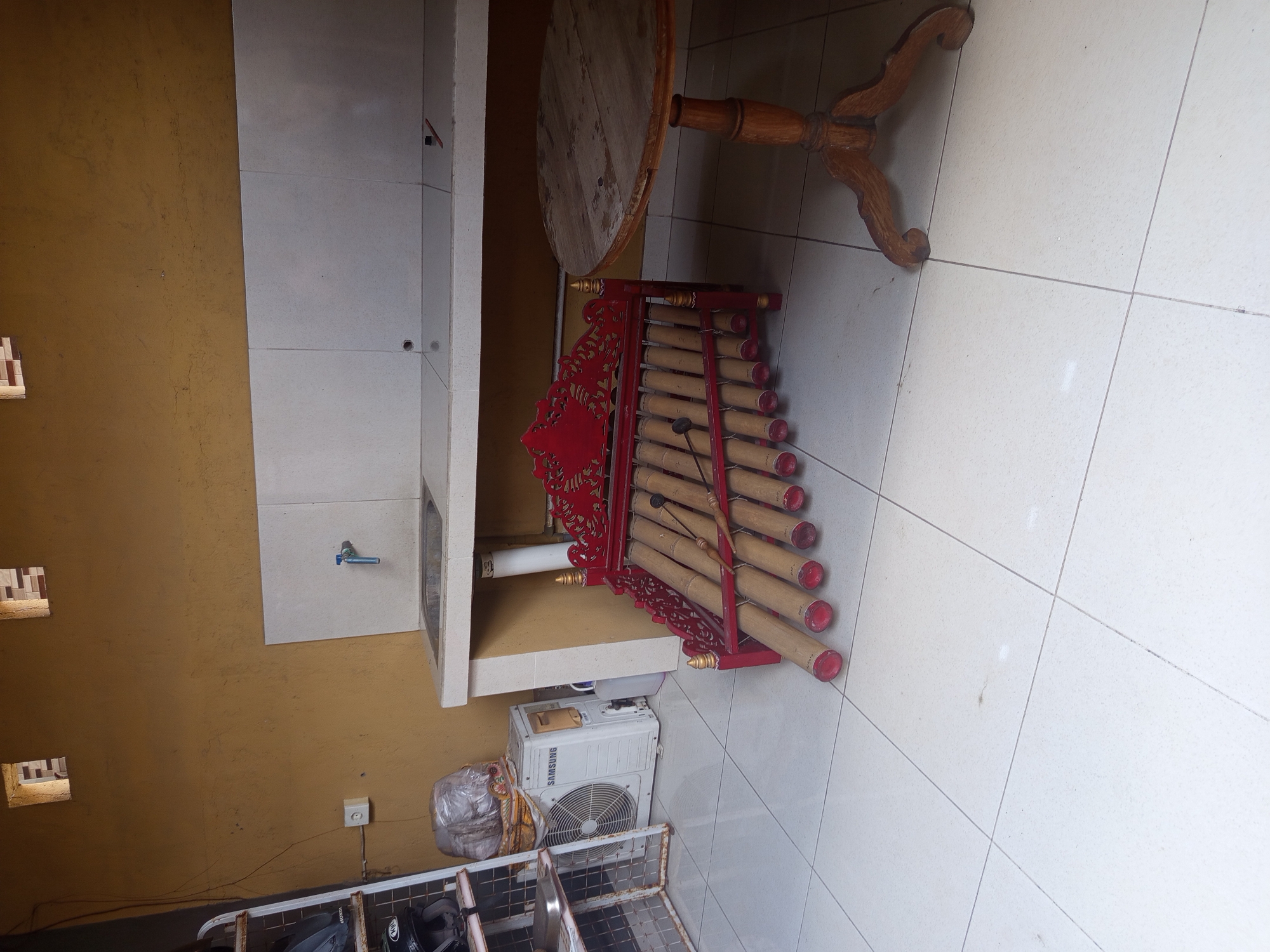 The image shows an indoor space in North Kuta, Indonesia. The floor is tiled with light-colored square tiles. A kitchen area is visible with light-colored cabinets and a sink. Below the cabinet is a Samsung air conditioner unit. To the left of the cabinets is a wall painted a yellowish-brown, with small square ventilation openings near the top. There is an electrical outlet on the wall. An angklung, a traditional Indonesian musical instrument made of bamboo tubes in a red frame, stands on the tiled floor near a small round wooden table with a decorative base. A rack or shelving unit with miscellaneous items is partially visible in the foreground. No people are visible in the image.