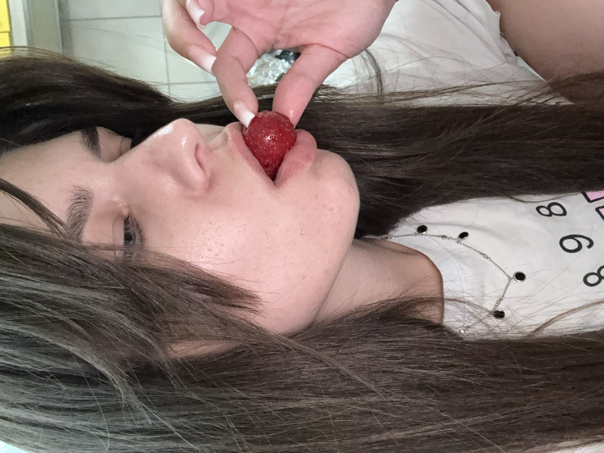 A close-up, profile shot shows a young woman with long, dark brown hair and fair skin. She is wearing a white t-shirt with a silver necklace featuring black circular pendants. Her right hand, with long, manicured white nails, is holding a single red strawberry to her lips. The background is blurred, suggesting an indoor setting with white tiled walls and possibly kitchen appliances. The lighting is soft and natural, highlighting the textures of her skin and hair.
