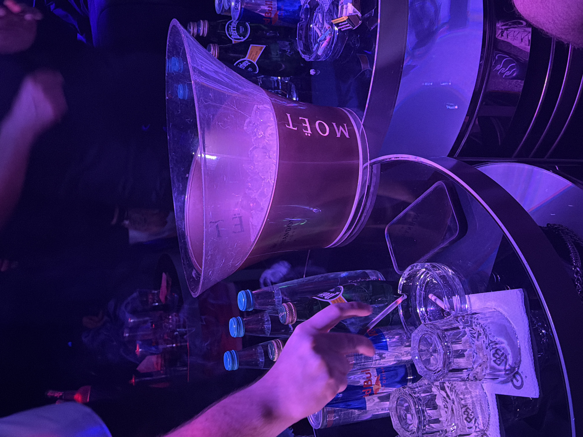 A person's hand reaches for bottles of water and Red Bull on a mirrored surface. Above them, an ice bucket with the word "MOËT" is visible. Other objects include glasses and ashtrays. The location is a dimly lit bar in Plovdiv, Bulgaria.