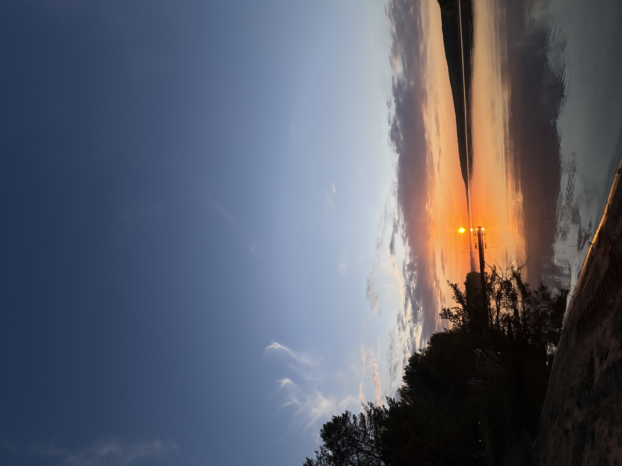 The image depicts a sunset scene over a body of water, likely a river or lake. The sky transitions from a deep blue on the left to orange and pink hues near the horizon. Clouds are present, reflecting the colors of the setting sun. The water reflects the sky and clouds, creating a mirror-like effect. Silhouetted trees and vegetation are visible in the foreground, obscuring part of the view. A lamp post or structure is visible in the center of the image, close to the sunset glow. Given the prompt, this scene is possibly located in Chaykovsky, Russia.