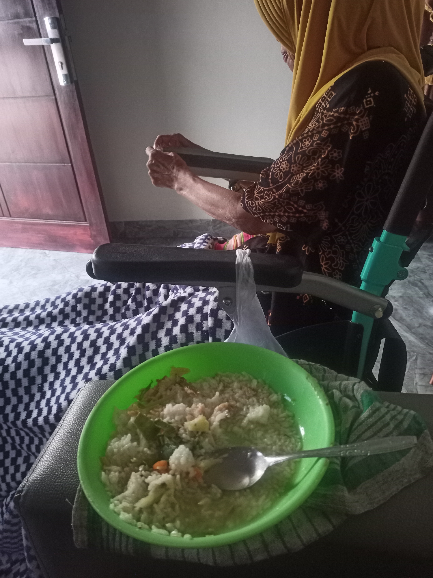 In Karangtalun, Indonesia, an elderly person in a wheelchair is presented with a meal in a green bowl, placed on a black surface covered with a checkered fabric. The individual, wearing a yellow hijab and a patterned brown and black long-sleeved shirt, is seen with their hands engaged with the wheelchair's armrest. The scene is set indoors, near a closed wooden door with a silver handle. The background consists of a light-colored wall and floor, suggesting a residential or care facility environment. The meal appears to be rice with some vegetables, and a spoon rests in the bowl. The lighting suggests it is daytime, with natural light entering the room.