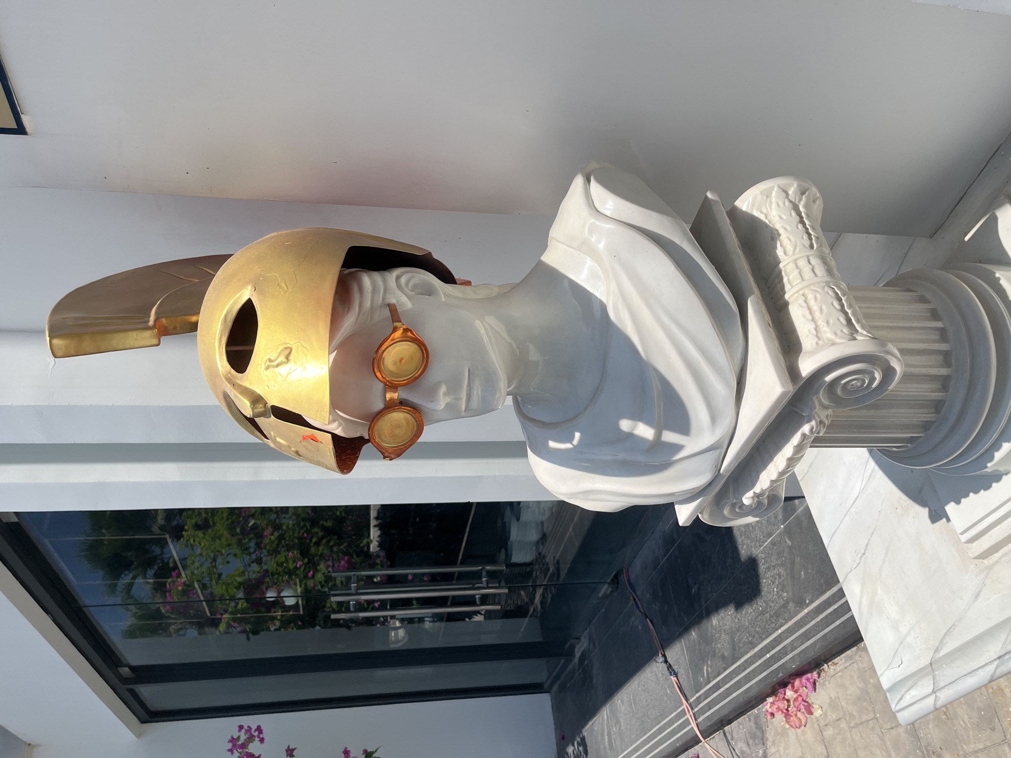 The image shows a white marble bust of a man adorned with a golden helmet and orange goggles, placed on a white marble pedestal. The bust is situated outdoors, in front of a building with large glass doors, reflecting trees and pink flowers. The pedestal is decorated with Corinthian columns. The scene appears to be in a courtyard or garden area, with shadows cast by the bright sunlight. The overall impression is one of a decorative art installation in a tropical setting. The specific location in Nha Trang, Vietnam, cannot be determined from the visual information provided.