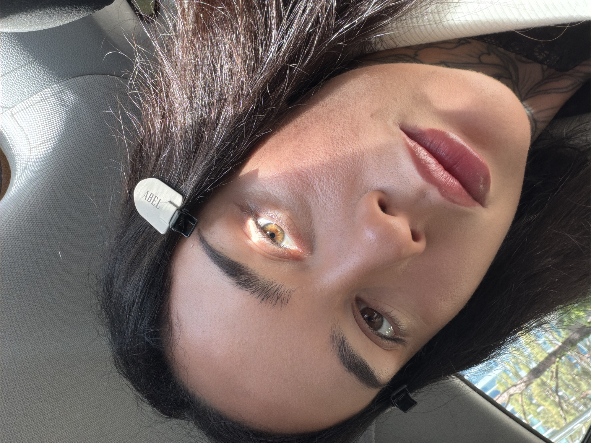 A close-up shot of a person's face tilted slightly to the side. The person has dark hair with clips holding it back, fair skin, and full lips with lip color. One eye is illuminated by a strong light source, revealing an amber color. The other eye is visible with a dark brown iris. A portion of a white garment with black lace details can be seen at the top right, suggesting the person may be wearing clothing. The background is blurred, showing hints of green foliage and a building suggesting an outdoor environment. The presence of a car interior (visible on the top-left) suggests the image was taken inside a vehicle. The location cannot be confirmed without more information.
