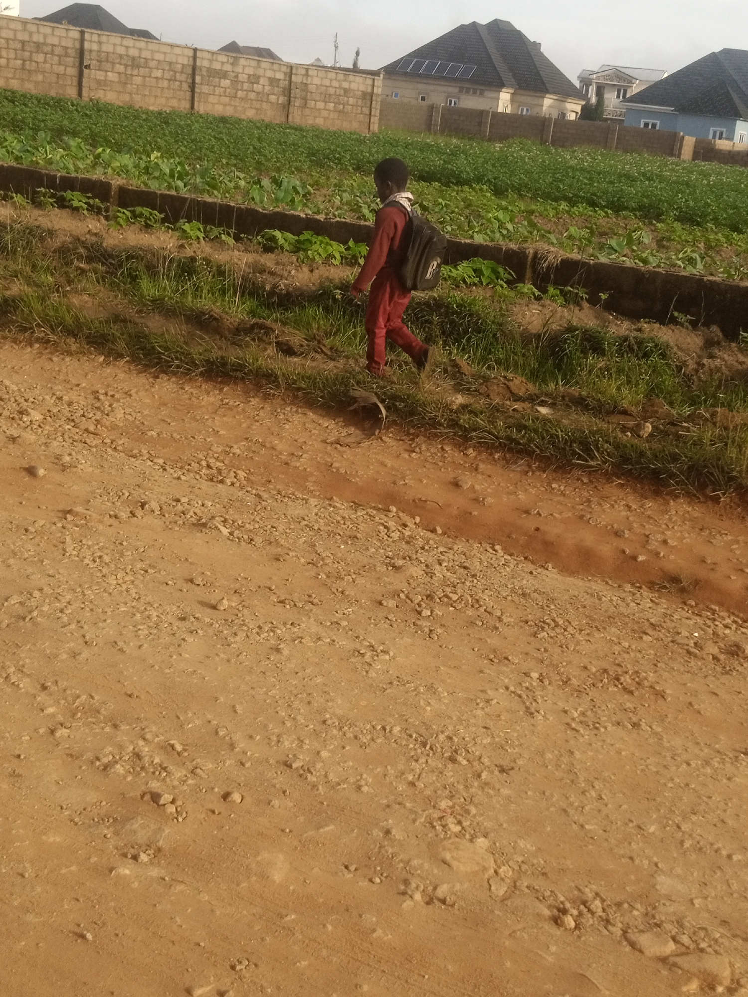 A child with a backpack walks on a dirt road in front of a cultivated field. The child is wearing a red outfit. In the background, there is a wall, houses, and a cloudy sky. The location is in the city of Russo, Nigeria.