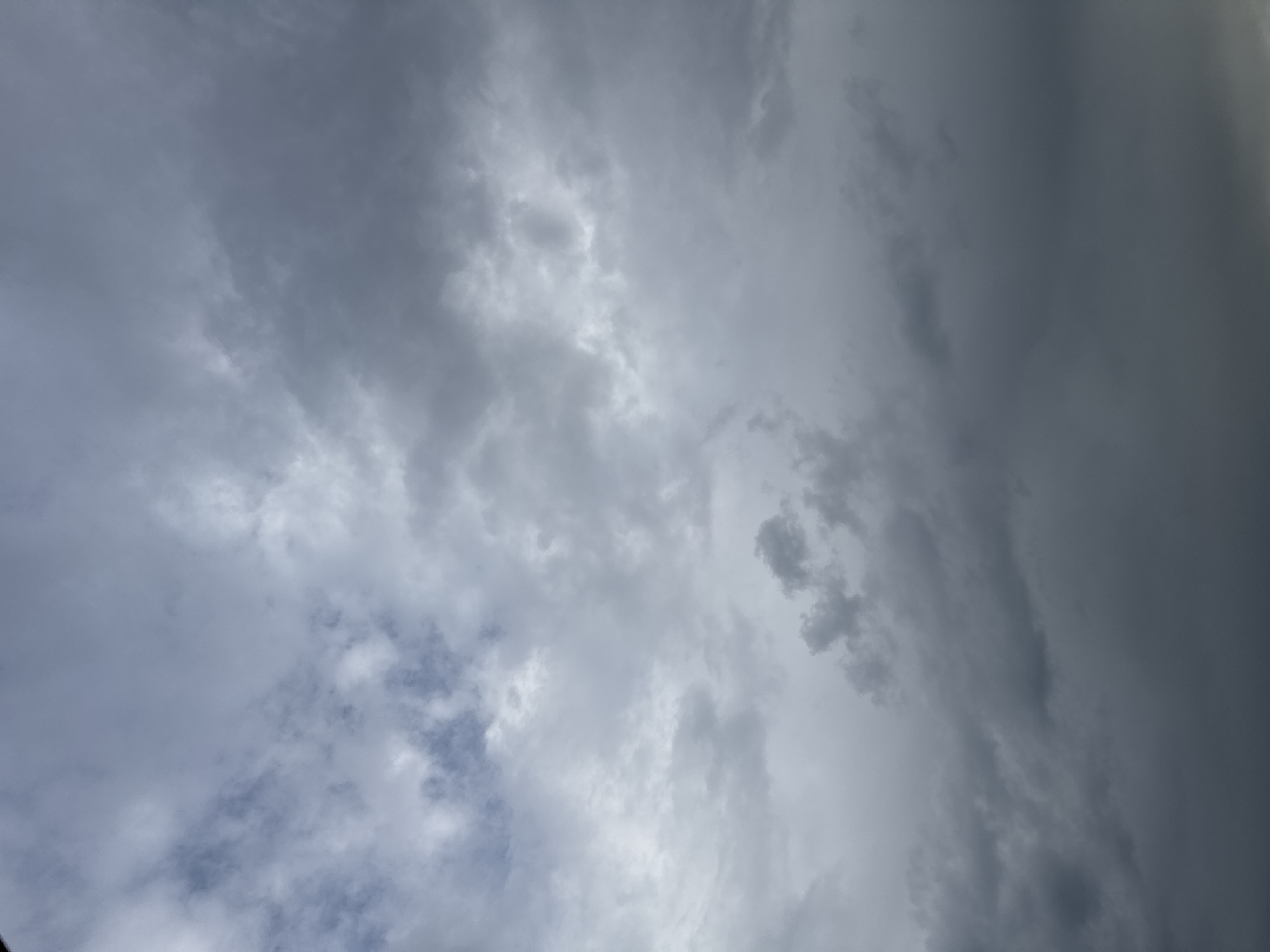 The image is a close-up, low-angle shot of a cloudy sky. The clouds are varied in shade and texture, ranging from light, wispy formations to dark, heavy masses that suggest impending rain. Some patches of lighter blue sky are visible through thinner clouds in the upper left quadrant. The lighting is diffused and overcast, creating a muted color palette dominated by grays and whites. There are no discernible landmarks or objects within the frame, suggesting the photograph was taken outdoors, looking directly upwards.