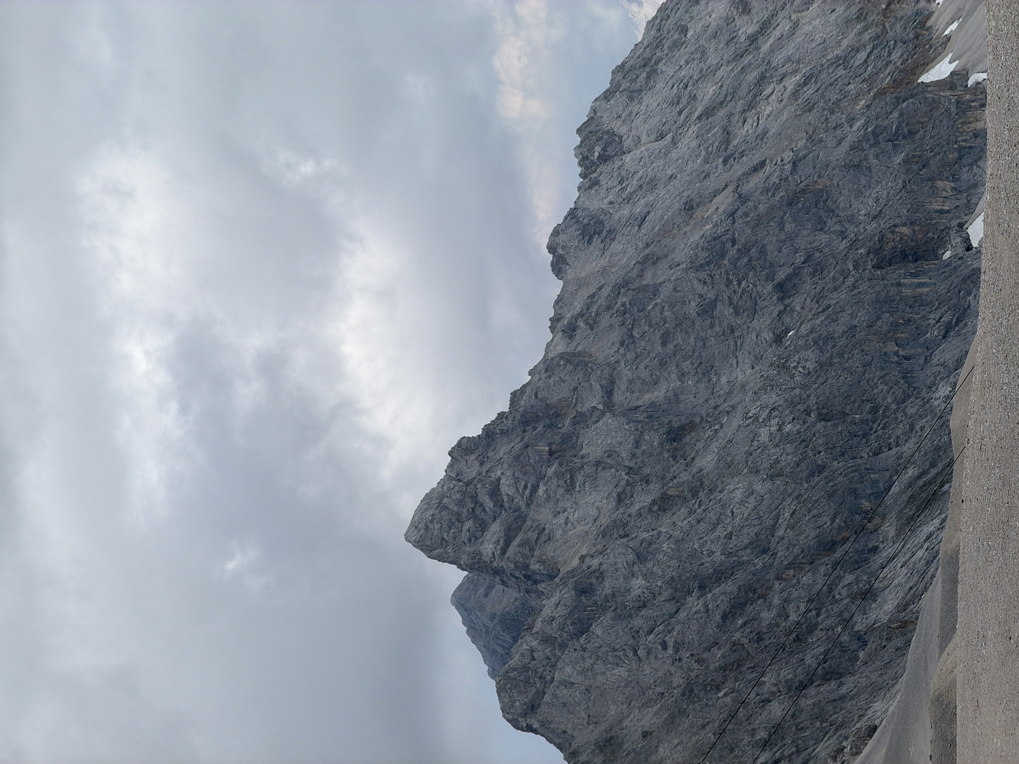 The image displays a majestic, rugged mountain range under a cloudy sky. The dominant subject is the imposing grey rock face of the mountain, characterized by its textured surface with numerous crevices and sharp peaks. In the foreground, a gravelly slope leads up to the mountain. The sky is overcast with varying shades of grey clouds, suggesting a cool or possibly impending weather change. There are no people or discernible structures visible in the image. The scene conveys a sense of natural grandeur and a remote, possibly alpine, environment. This photograph is likely of the Zugspitze massif, the highest peak in Germany, located near Garmisch-Partenkirchen.