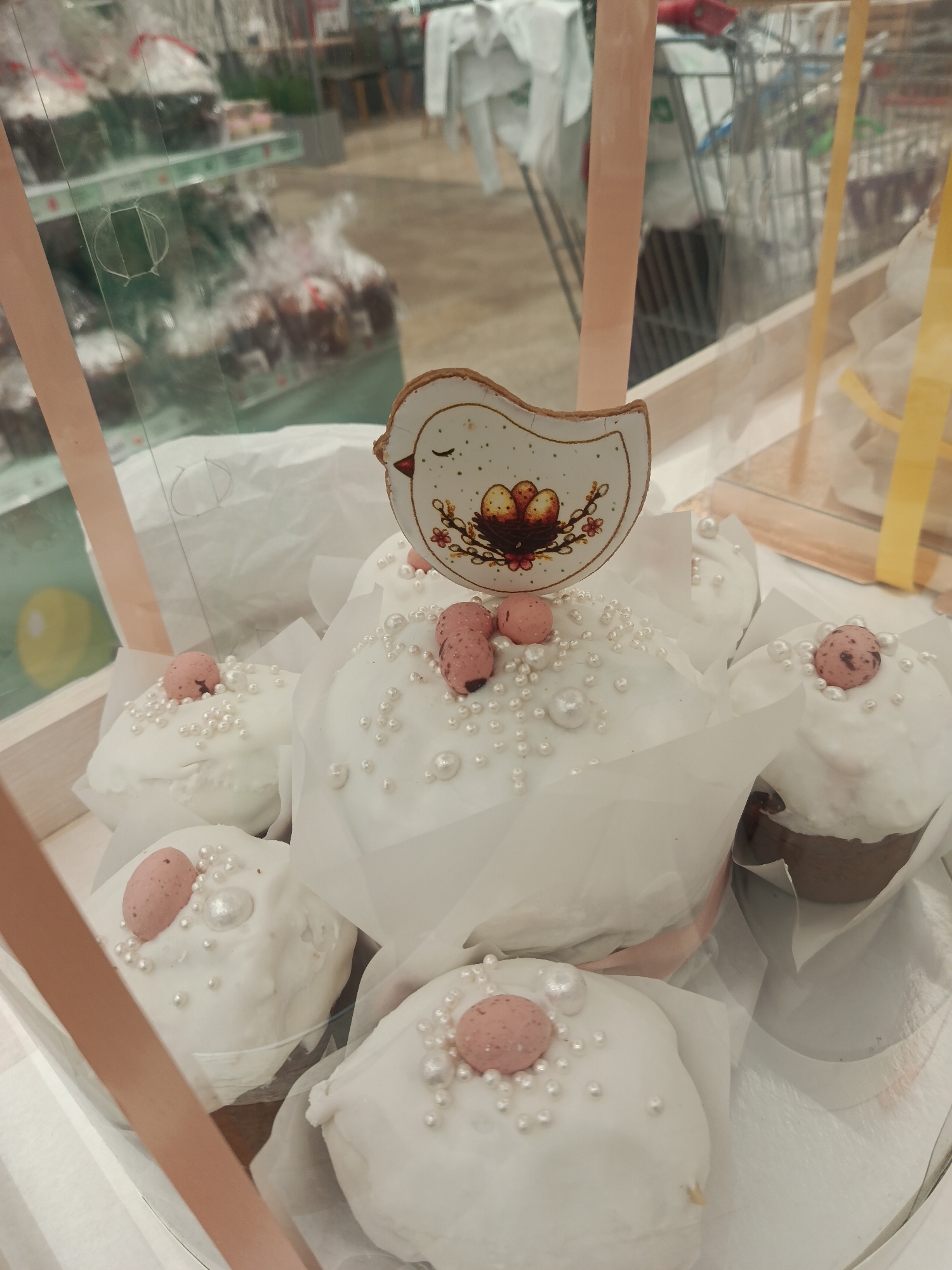 The image displays a close-up view of several beautifully decorated individual cakes, likely Easter kulichi, presented within a transparent display box or packaging. Each cake is generously covered with a smooth white icing, embellished with an assortment of small, round, pearlescent sprinkles. Many of the cakes also feature a single, speckled pink, egg-shaped candy placed on top, adding a thematic touch.
A prominent decorative element is a bird-shaped topper, possibly made of wood or ceramic, resting on the largest central cake. This topper is white with a simplified depiction of a bird with a closed eye and features a colorful illustration of a nest containing three speckled eggs, surrounded by small branches with pink flowers. The cakes are nestled in white paper wrappers, suggesting they are prepared for retail.
The background is blurred but reveals a retail environment, likely a supermarket or bakery. Reflections and distortions on the clear display case show shelves stocked with items, some wrapped in plastic, and further back, the recognizable forms of shopping carts. The overall aesthetic suggests a festive, springtime celebration, strongly indicative of Easter preparations, aligning with the context of Moscow, Russia, where decorated kulichi are a traditional part of the holiday. There is no discernible text visible in the image.