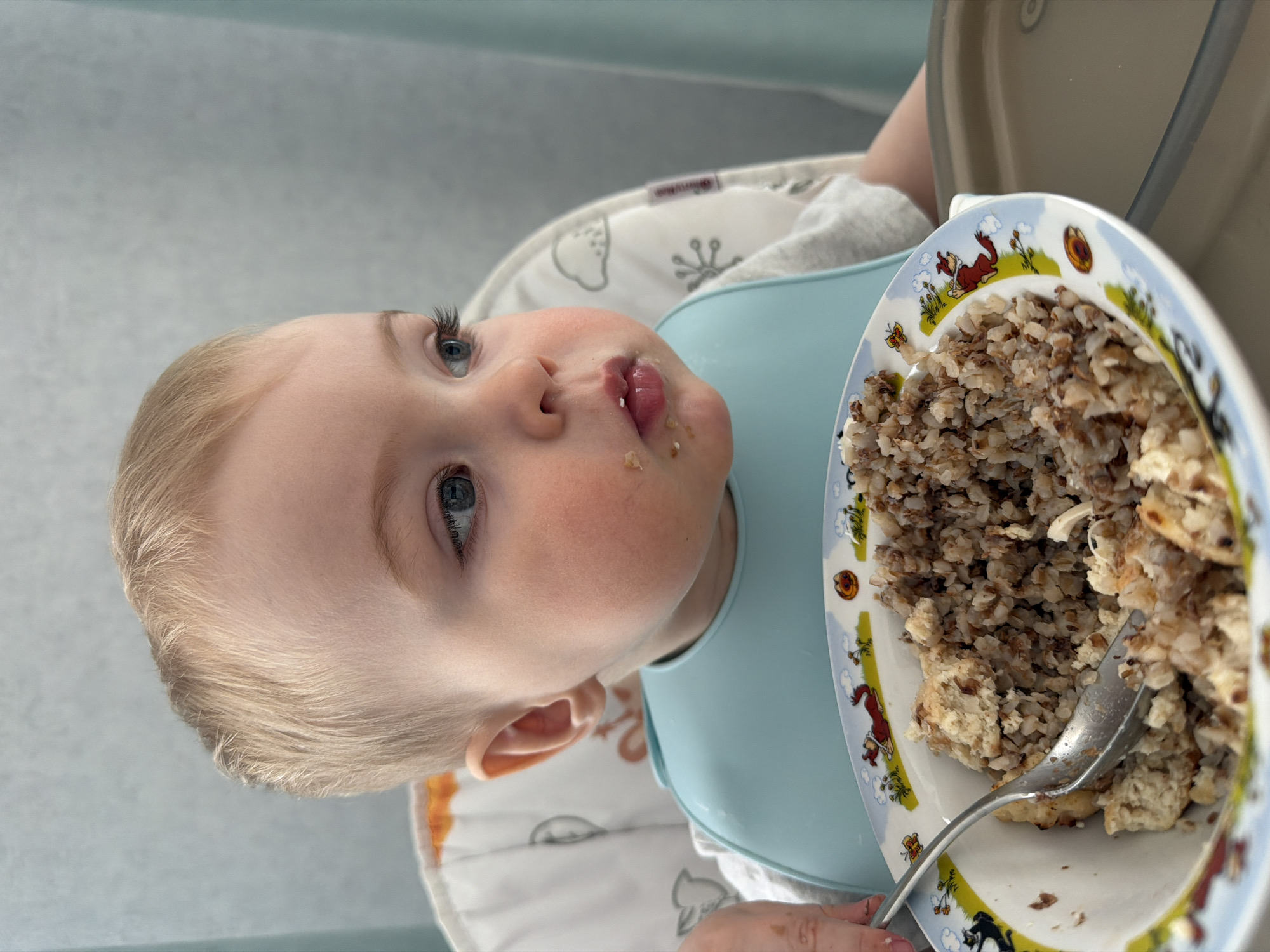 A baby with blonde hair and blue eyes sits in a high chair, wearing a light blue bib. Food residue is visible around the baby's mouth. The baby looks up and slightly to the left. A plate of buckwheat and another food sits in front of the baby. A silver spoon rests in the plate of food. The plate is decorated with cartoon animals. The high chair has a white and gray patterned seat. The background is a light gray. There is no information to confirm the location is Votkinsk, Russia.