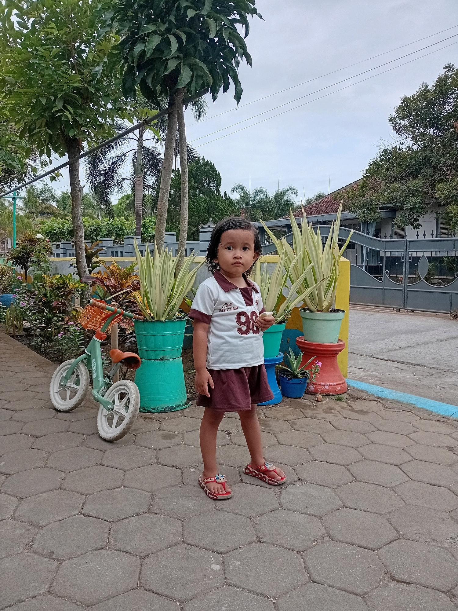 A young girl stands in the foreground on a hexagonal paved walkway in Gebang, Indonesia. She is wearing a white polo shirt with a maroon collar and trim, featuring the number "98" and text that reads "CHILL TAKE IT EASY." She also wears a maroon skirt and red flip-flops. To her left, a teal balance bike with a basket is parked. Behind her, several potted plants are arranged, including large, spiky plants in turquoise pots and a smaller plant in a blue pot, and an orange pedestal with a small plant and pink flower. The background reveals a grey metal gate, a house with a red tile roof, and lush greenery including trees and palms. The sky is overcast.