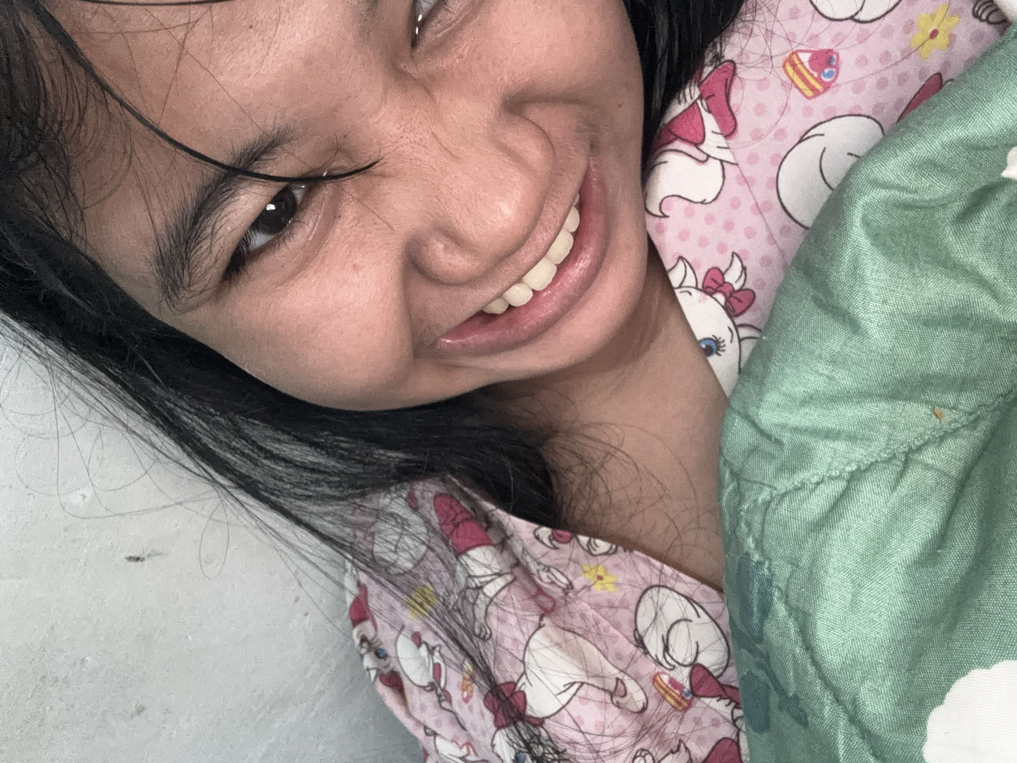 A close-up, overhead shot captures a young individual with dark hair and fair skin. They are lying down, with their head resting on a green textured blanket. Their mouth is open in a wide smile, revealing teeth. The individual is wearing a pink pajama top adorned with a repeating pattern of cartoon characters, specifically white cats with pink bows. To the left of the individual, a white, slightly textured wall is visible. The image is a selfie, indicating it was taken by the subject. The specific location is in North Jakarta, Indonesia.