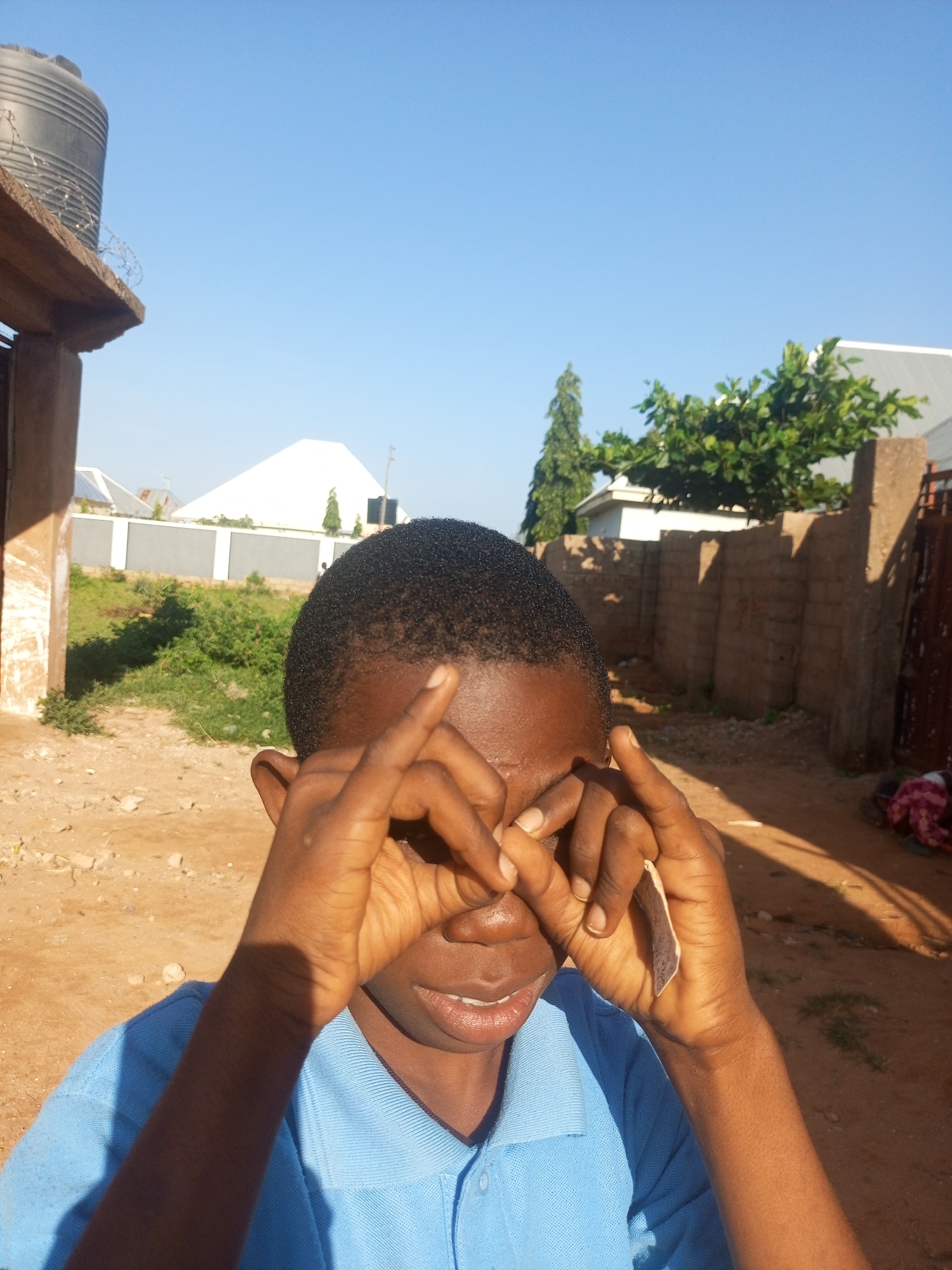 The image depicts a young Black male in the foreground, wearing a blue polo shirt. He is holding his hands up to his eyes, forming a circular shape with his fingers, as if peering through binoculars or a makeshift lens. His facial expression is difficult to discern clearly due to the way his hands obscure his eyes. The background reveals an outdoor environment characterized by a dusty, dirt ground. In the middle ground, there is a low, weathered brick wall and sparse green vegetation. Beyond the wall, a few buildings are visible, including one with a white, angled roof and another with a darker, possibly corrugated metal roof. A tall, thin tree stands to the left of the white-roofed building, and a larger, leafy tree is positioned further to the right, behind the brick wall. The sky above is clear and blue. A dark gray water tank is mounted on a concrete structure to the far left, partially obscured by the edge of a building in the immediate foreground. The scene appears to be set in a semi-arid or rural area within a city, such as Tempure, Nigeria, given the architectural styles and vegetation.
