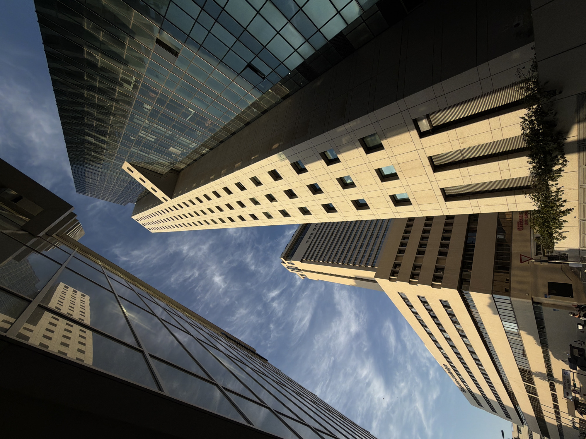 The image depicts a low-angle perspective of a cluster of modern buildings against a clear blue sky with scattered wispy clouds. The architecture is dominated by glass facades and concrete structures, characteristic of urban environments.
One prominent building, on the right side of the frame, is a tall, angular skyscraper with a dark glass exterior that reflects the sky and surrounding structures. A lighter-colored, stepped building is partially visible in front of it, with numerous small square windows.
To the left, another building features a glass facade with a geometric pattern of diagonal lines. Its reflection of another building with a grid of windows is clearly visible in the glass.
In the background, a long, beige-colored building with many horizontal windows stretches across the middle ground. The top of this building displays text in Hebrew, which reads "מגדל אמות השקעות" (Migdal Amot Investments).
The overall impression is one of a modern cityscape, likely in a business district. The lighting suggests it is daytime, with shadows cast by the buildings indicating the sun's position. There are no visible people or specific actions occurring that would suggest a particular event. The scene is static and architectural.