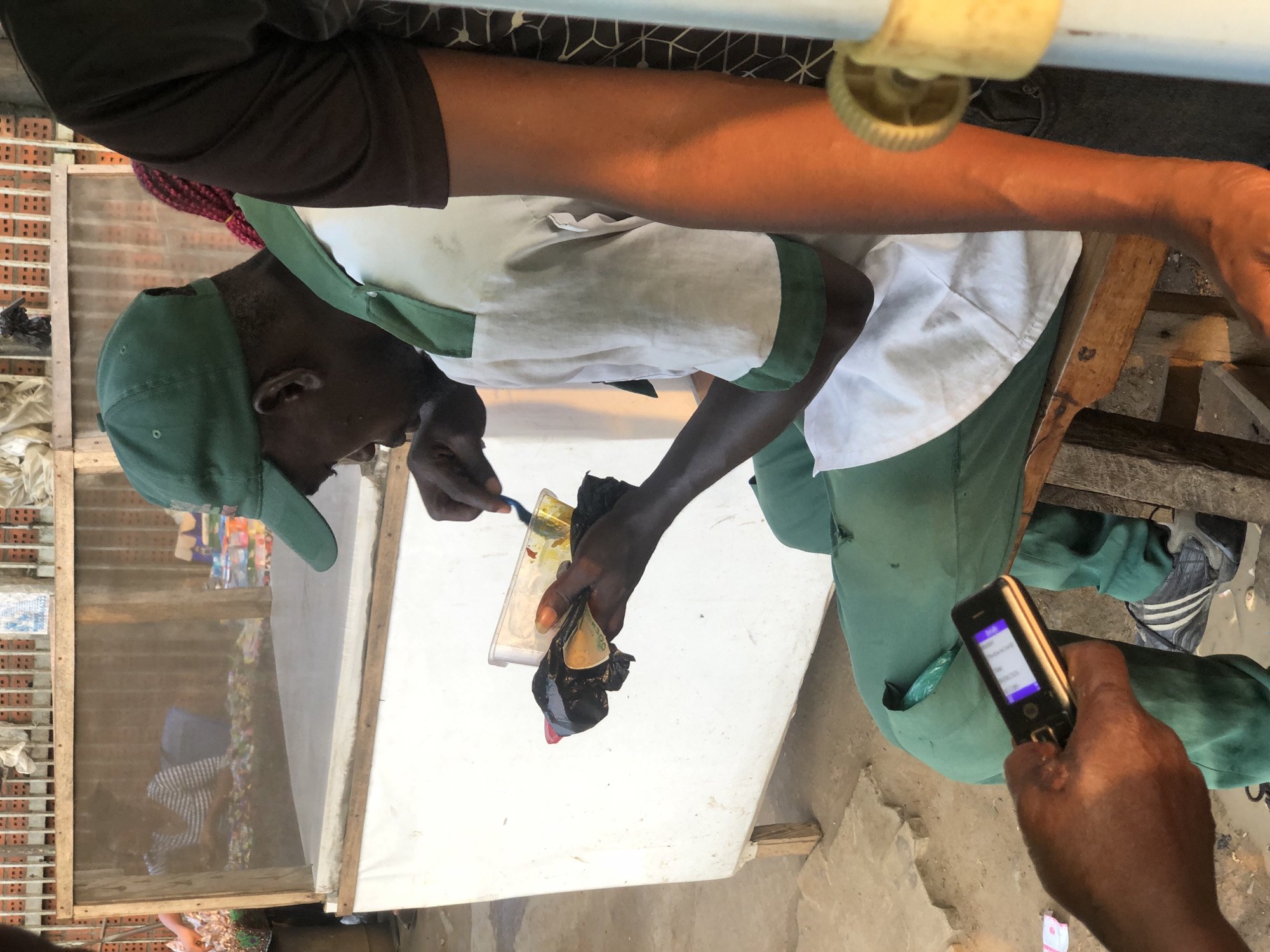 In Ikeja, Nigeria, an individual wearing a green cap and a white and green uniform is seated on a wooden stool. They are holding a clear plastic container with food and a blue utensil, appearing to be in the process of eating or preparing food. A black plastic bag is also held in their hands. Nearby, another person's hand is visible holding a flip phone. In the background, a stall structure with a white front and wooden frame is present, with a mesh screen. The ground is dirt and concrete. The overall scene suggests an outdoor market or street-side food vendor environment.