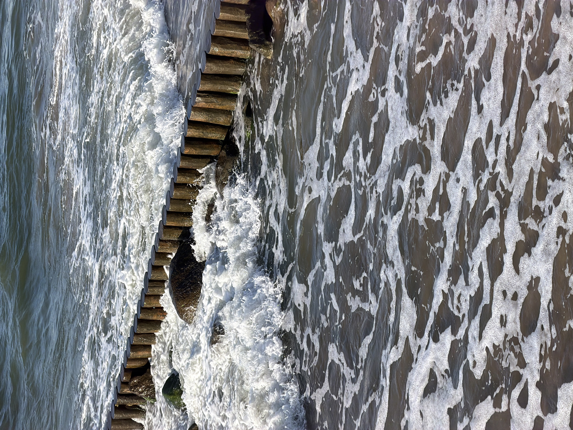 A view of the Baltic Sea coastline in Zelenogradsk, Russia, shows wooden groynes, rocks, and waves breaking. The groynes are made of vertical wooden posts driven into the seabed, arranged in a parallel line. The water is dark and turbulent, with white foam and spray generated by the breaking waves. Several dark-colored rocks are visible among the groynes, some partially submerged by the water. The sky is overcast, and the overall lighting suggests a cloudy day. The scene captures the dynamic interplay of water and man-made structures on a beach.