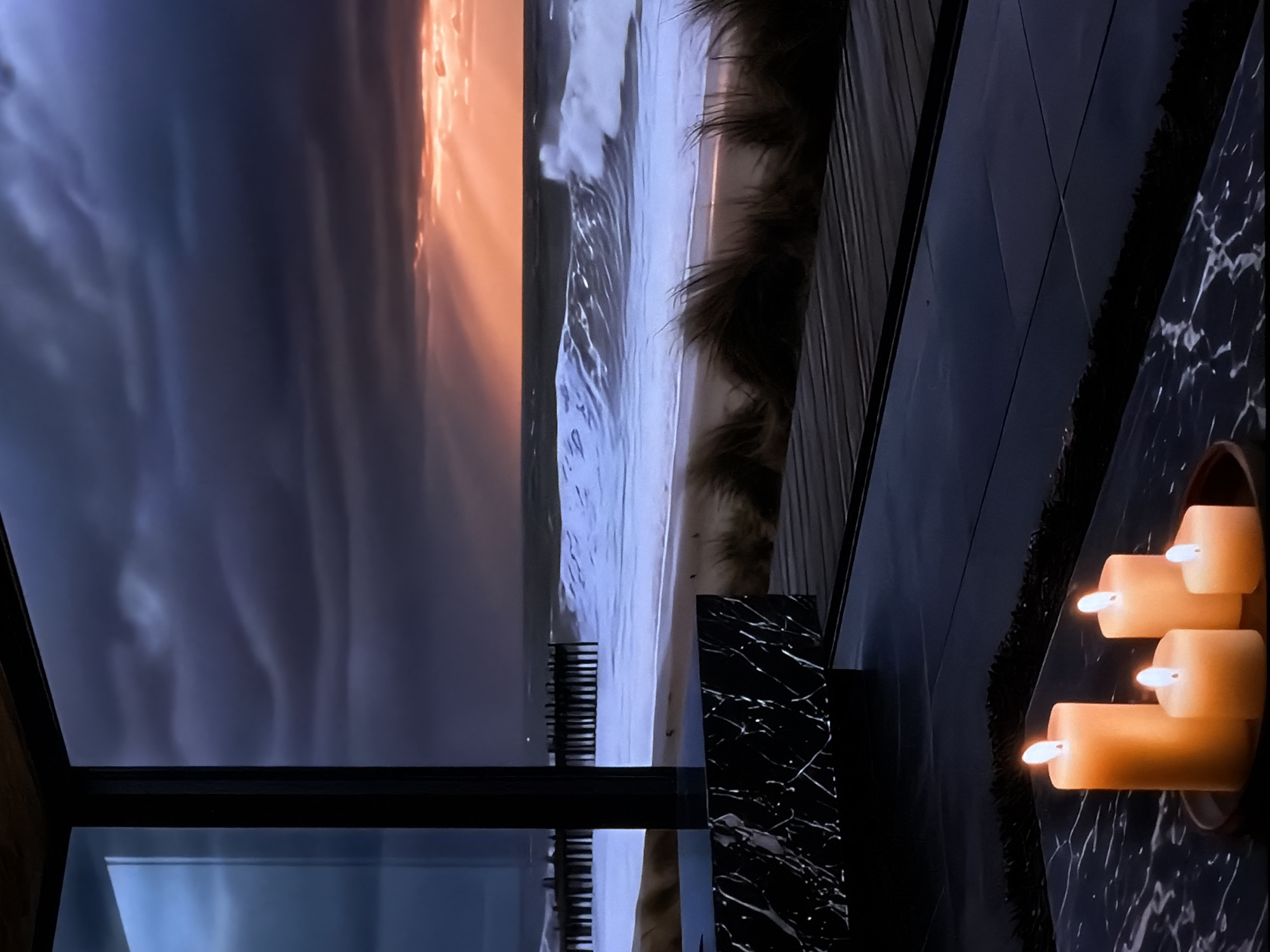 This image captures a dramatic coastal sunset viewed from a modern interior. The scene is divided by a dark window frame, offering a glimpse into both the tumultuous outdoors and a cozy indoor setting.
The outdoor portion reveals a dynamic seascape at dusk. The sky transitions from dark, stormy grey clouds at the top to a vibrant, fiery orange at the horizon, where the setting sun casts luminous rays through the clouds. Powerful white-capped waves crash energetically onto a sandy beach, creating intricate foamy patterns. Several dark, vertical structures, likely wooden groynes or breakwaters, extend into the turbulent water. Beyond the beach, a stretch of dark, shaggy dune grass is visible.
The indoor foreground features a sophisticated aesthetic. A sleek, black surface with distinctive white marble veining serves as a prominent element. On this surface, a cluster of four cylindrical candles, appearing to be electric or LED with steady, warm glows, are arranged in a circular holder, emitting a soft, inviting light. A dark, textured element, possibly a decorative rug or upholstery, is visible next to the candles, complementing the dark interior design. The window frame is dark and minimalistic, framing the expansive view.
The time of day is sunset or dusk, evidenced by the brilliant sky and the lit candles. The weather appears dramatic, with a stormy sky and strong waves. The scene conveys a sense of warmth and contemplation juxtaposed against the raw power of nature. No visible text is present. While the provided location context is Sofiivska Borshchahivka, Ukraine, the depicted scene is clearly a dramatic ocean or sea coast, suggesting the photo's origin rather than its subject matter.