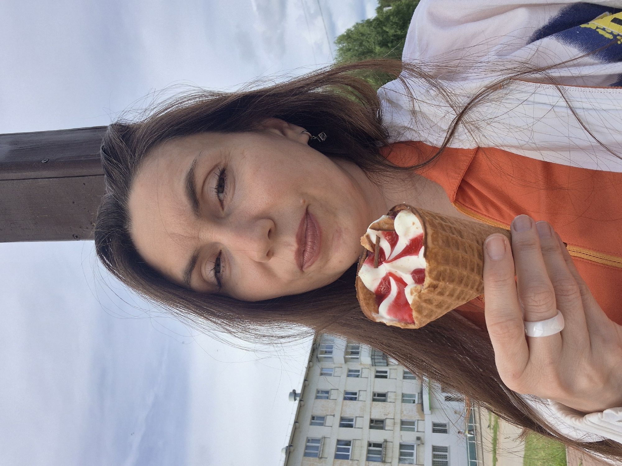 The image displays a close-up, rotated portrait of a woman holding an ice cream cone. She has long, dark brown hair cascading around her face and shoulder. Her expression is calm, with her eyes partially closed. She wears a white top or jacket over an orange garment, with a subtle blue and yellow pattern visible on the white fabric. A simple white ring is on her right ring finger, and a small, square-shaped earring adorns her left ear.
She holds a waffle cone filled with creamy white ice cream, streaked with a vibrant red fruit swirl, likely strawberry or cherry, and topped with thin chocolate sticks.
The background is an outdoor scene under a cloudy sky. A multi-story, light-colored building with many windows occupies the lower half of the frame (due to the image rotation), suggesting an urban or suburban setting. Green foliage from trees is visible on the right side, and a dark wooden post or beam is present on the far left. The natural light indicates it is daytime. There is no visible text in the image.