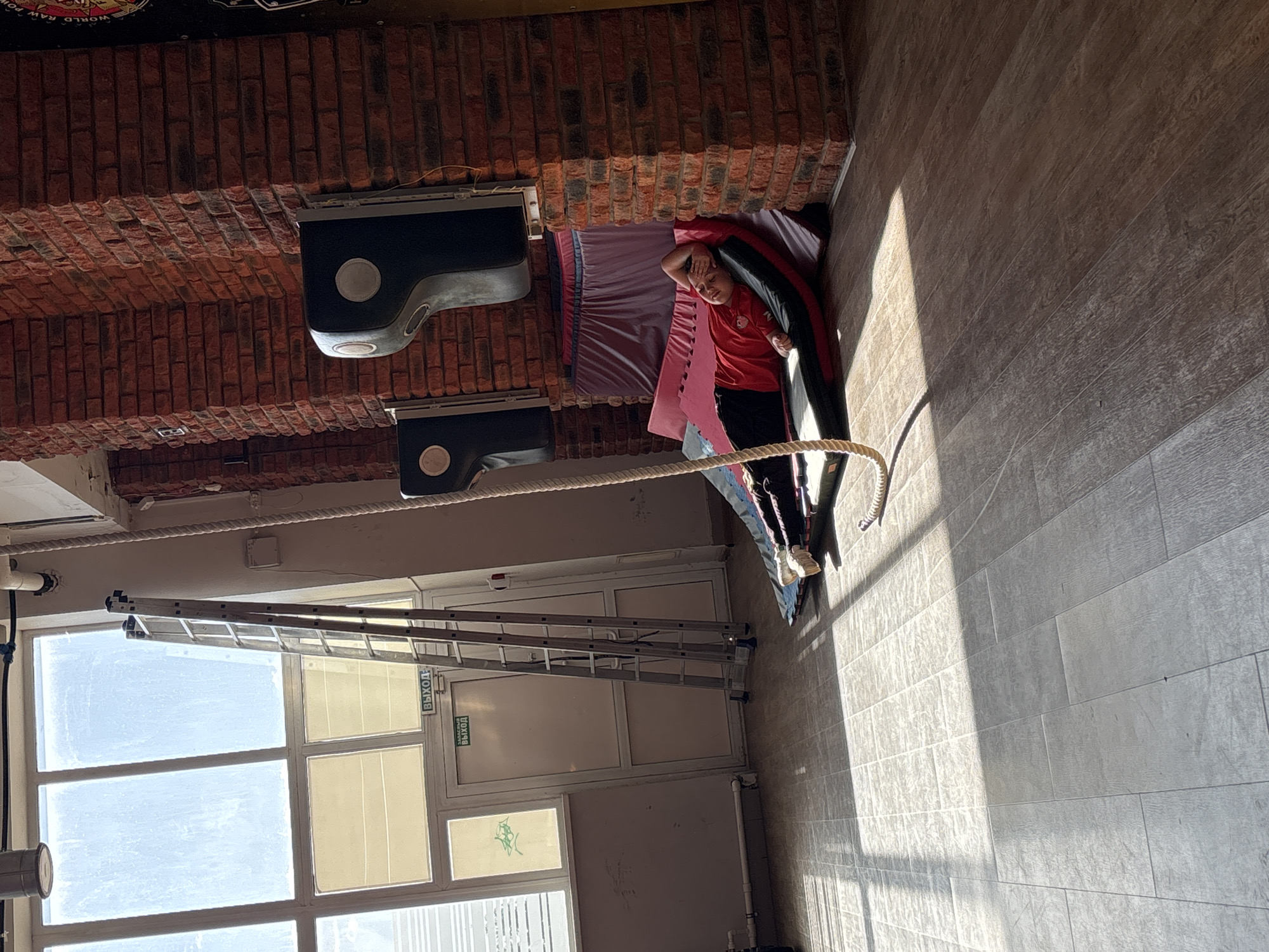 A person in a red shirt and black pants lies on a mat on the floor in a room with brick walls. They are resting their head on their hand. A rope is stretched across the room near them. A ladder is leaning against a wall with windows and a door. The door has a sign in Russian. Two black objects are attached to the brick wall. The floor is tiled. The location is the city of Chaikovsky, Russia.
