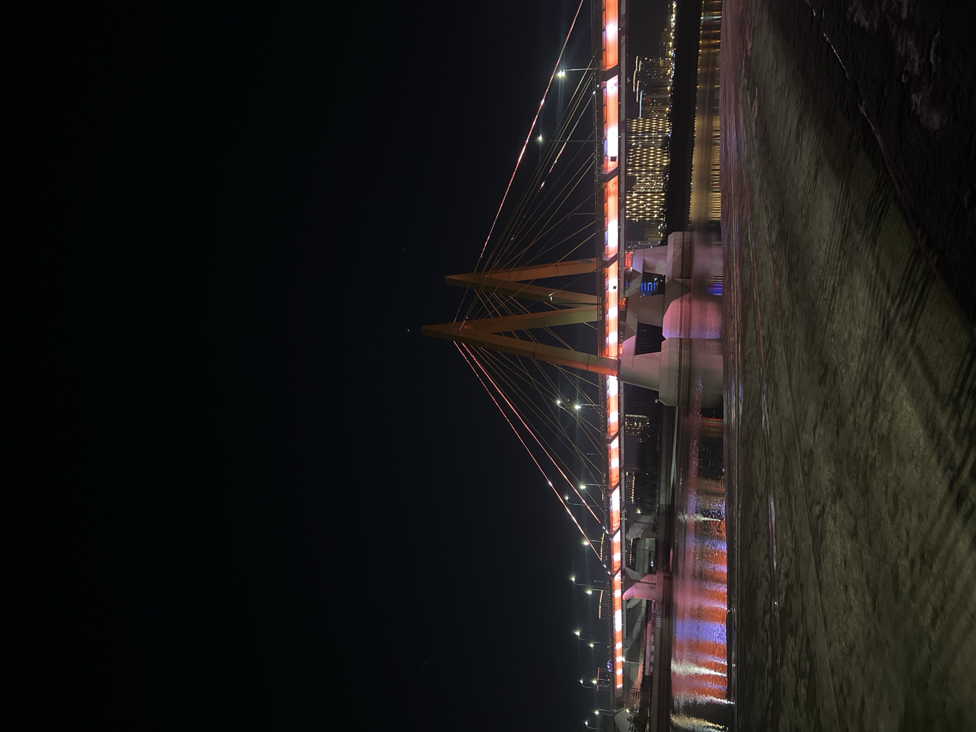 The image shows a brightly lit bridge at night. The bridge has an inverted Y-shaped support structure at the top, with cables extending downwards from the support to the road surface. The support and cables are illuminated, and the road surface has lights along it. Reflections of these lights are visible in the nearby water. There are illuminated buildings in the background on the opposite side of the water. The overall lighting is bright, and the sky is black. The photograph is taken from a low angle. The image shows the Millennium Bridge in Kazan, Russia.