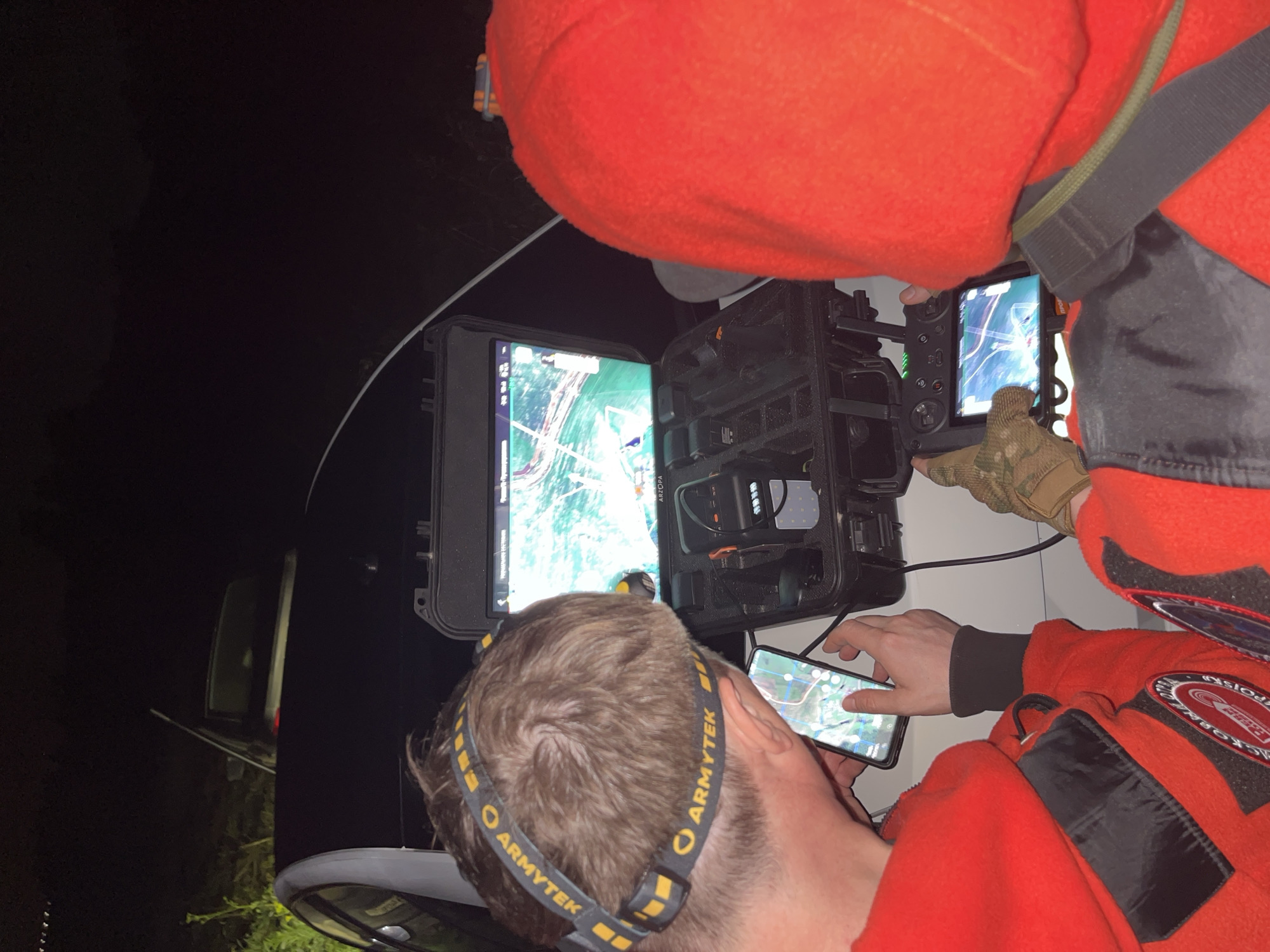 Two individuals, wearing matching red hooded jackets and headlamps, are engaged in operating a drone at night. One individual on the left is holding a smartphone displaying a map interface, while the other individual on the right is operating a drone controller with a screen. They are positioned next to a black vehicle, and a case containing drone equipment is open between them. The scene appears to be outdoors, with vegetation visible in the background, and is consistent with a location in or around the village of Badashka, Russia. The lighting suggests it is dark, with the headlamps and screens being primary light sources.