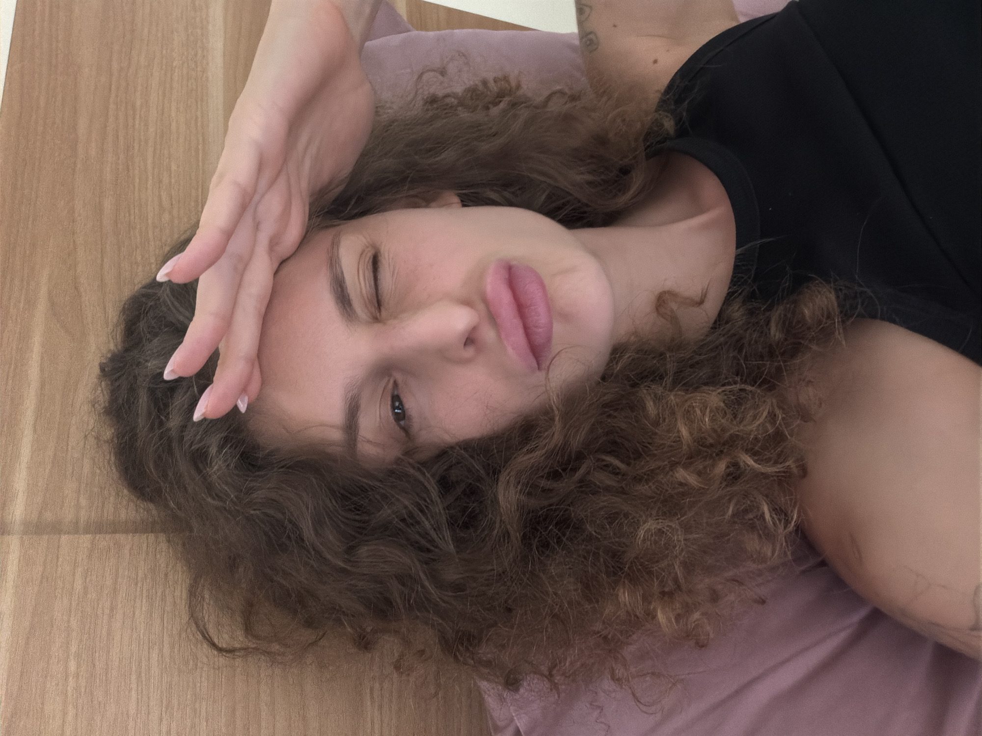 A young woman with long, curly dark brown hair is captured in a close-up, informal portrait, lying down and looking towards the camera. She appears to be reclining on a soft light purple surface, likely a pillow or bed, which occupies the right side of the frame. To the left, a warm-toned wooden surface, possibly floorboards or paneling, forms the background.
The woman's right hand is raised and gently rests on her forehead, with her fingers slightly splayed, revealing light-colored manicured nails. Her left eye is open, while her right eye is partially closed, creating a winking or squinting effect. Her lips are softly pursed in a pout. She is wearing a simple black t-shirt, and a subtle tattoo is visible on her upper right arm. The overall expression conveys a playful and relaxed mood. There is no visible text in the image. The setting suggests an indoor environment, potentially in North Kuta, Indonesia, as indicated by the location context.