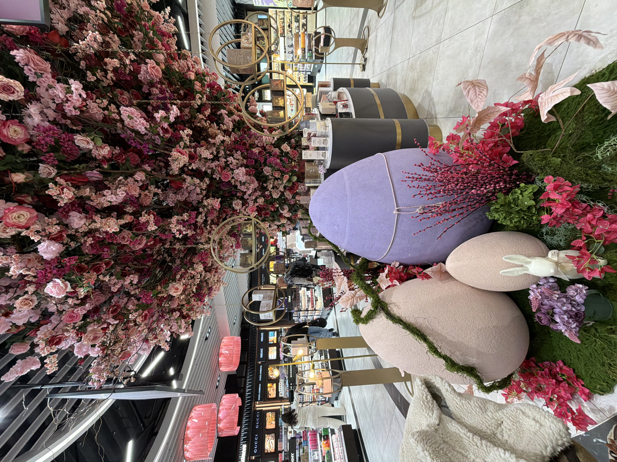 The image shows a store display, potentially a department store, in Sofia, Bulgaria. The focus is on a decorative installation featuring oversized Easter eggs in pastel colors (lavender and pink) adorned with artificial flowers, moss, and a small white bunny figurine. A large floral arrangement, primarily in shades of pink and red, hangs overhead. The display is located in a shopping area with shelves filled with cosmetic products and fragrances. Customers can be seen browsing the merchandise. The setting includes modern lighting fixtures, such as square and circular light fixtures, and polished floors, suggesting an upscale retail environment.