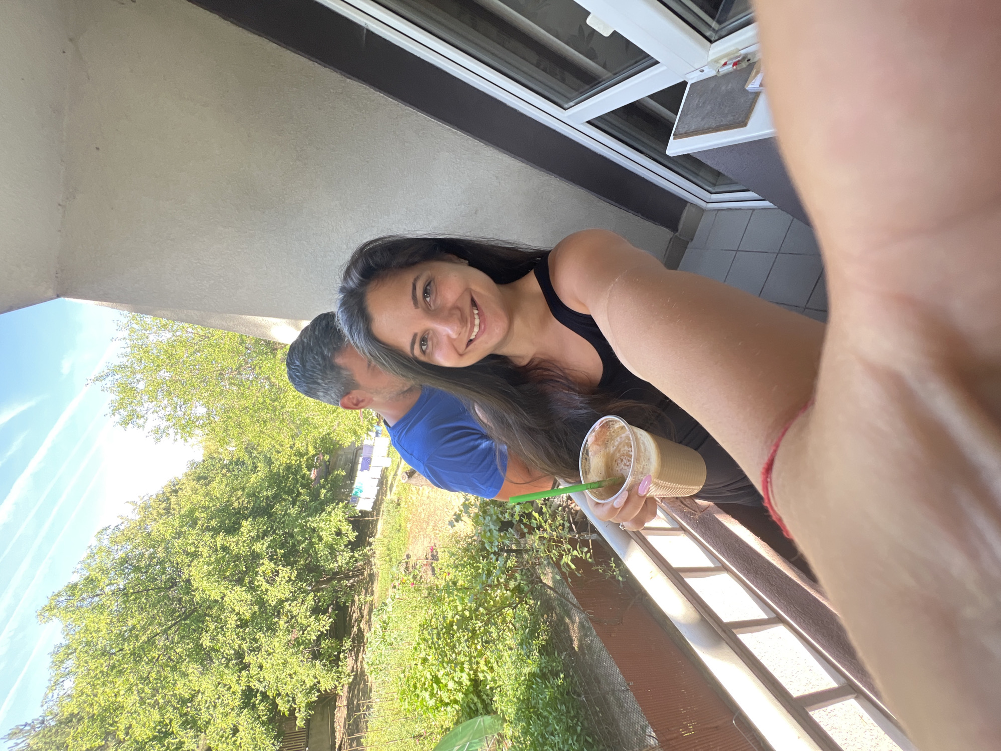 The image depicts a selfie taken by a woman with long, dark hair, wearing a black tank top. She is smiling and holding a disposable cup with a frothy brown beverage and a green straw in her right hand. Her left arm is extended towards the camera. Behind her, a man with short, dark hair and a blue t-shirt is partially visible. They are standing on a balcony with a metal railing. In the background, there is a garden with lush green plants, some laundry hanging on a line, trees, and a bright blue sky with white clouds. The scene appears to be set in a residential area, possibly in the city of Sofia, Bulgaria, given the context of the question.