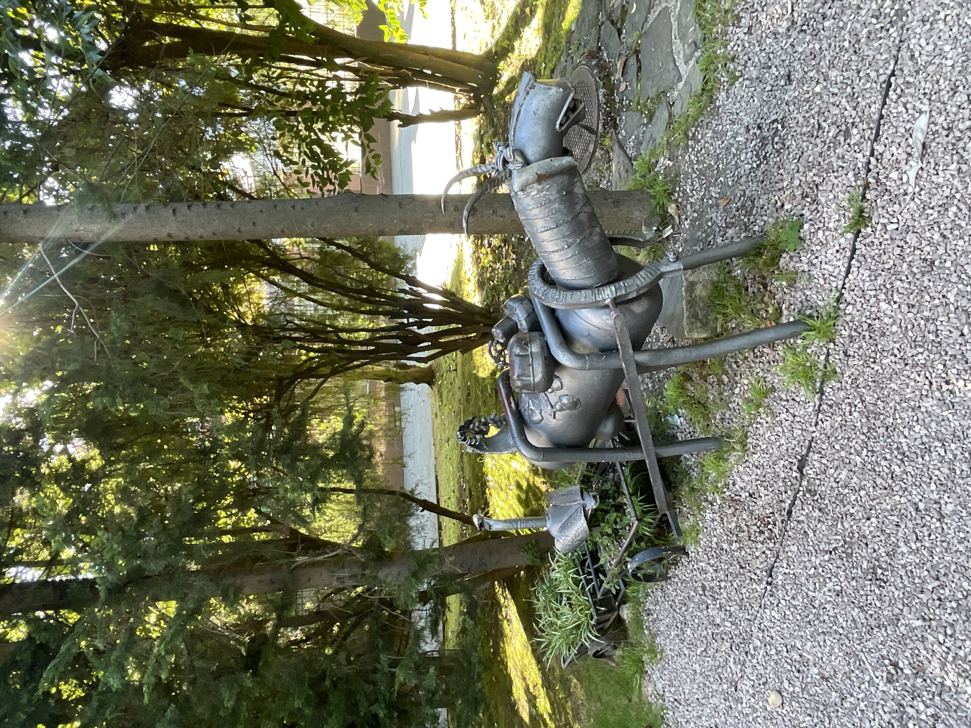 In the city of Sochi, Russia, a whimsical metal sculpture of a goat-like creature stands in a garden. The sculpture is made of salvaged metal and features a cylindrical body, legs, and a head with horns. A smaller cart filled with plants is attached to the front of the sculpture, with what appears to be a metal goose or swan head peeking out. The scene is set amidst lush green trees and dappled sunlight, with gravel paths and stone paving stones visible on the ground. The overall impression is one of rustic charm and imaginative artistry within a natural setting.