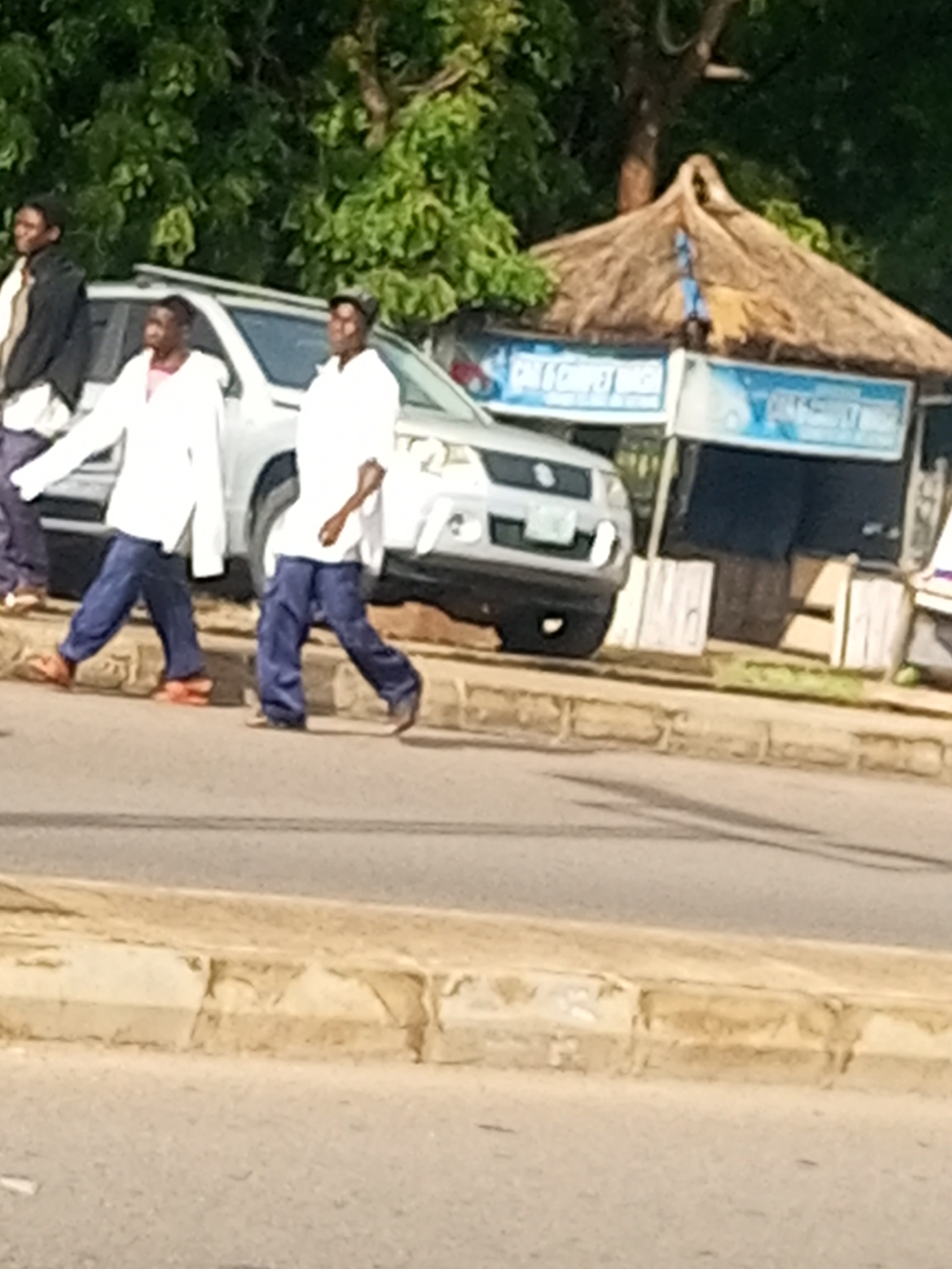 In Tempure, Nigeria, on a sunny day, three men are walking along the side of a road. The man on the far left is wearing a dark jacket and a light shirt, with dark pants. The man in the middle is wearing a white long-sleeved shirt and dark pants. The man on the right is wearing a white long-sleeved shirt and dark pants. They are walking past a silver SUV that is parked on the side of the road. Behind the SUV is a thatched-roof stall with blue signage. The ground is paved with asphalt and there is a concrete curb separating the road from a grassy area. The background is filled with lush green trees.