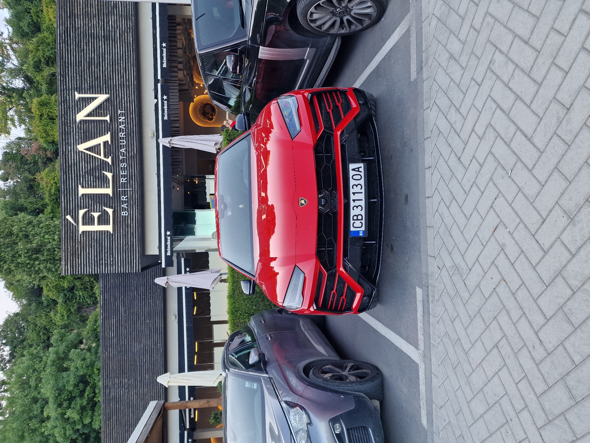 A red Lamborghini Urus is parked in a parking spot in front of a building with a sign that reads "ÉLAN BAR RESTAURANT." To the left of the Lamborghini is a gray Peugeot SUV, and to the right is a black Range Rover. The parking lot is paved with interlocking bricks. The building has a dark tiled roof and large windows with black awnings. There are trees in the background. The license plate on the Lamborghini reads "CB 3113 OA."