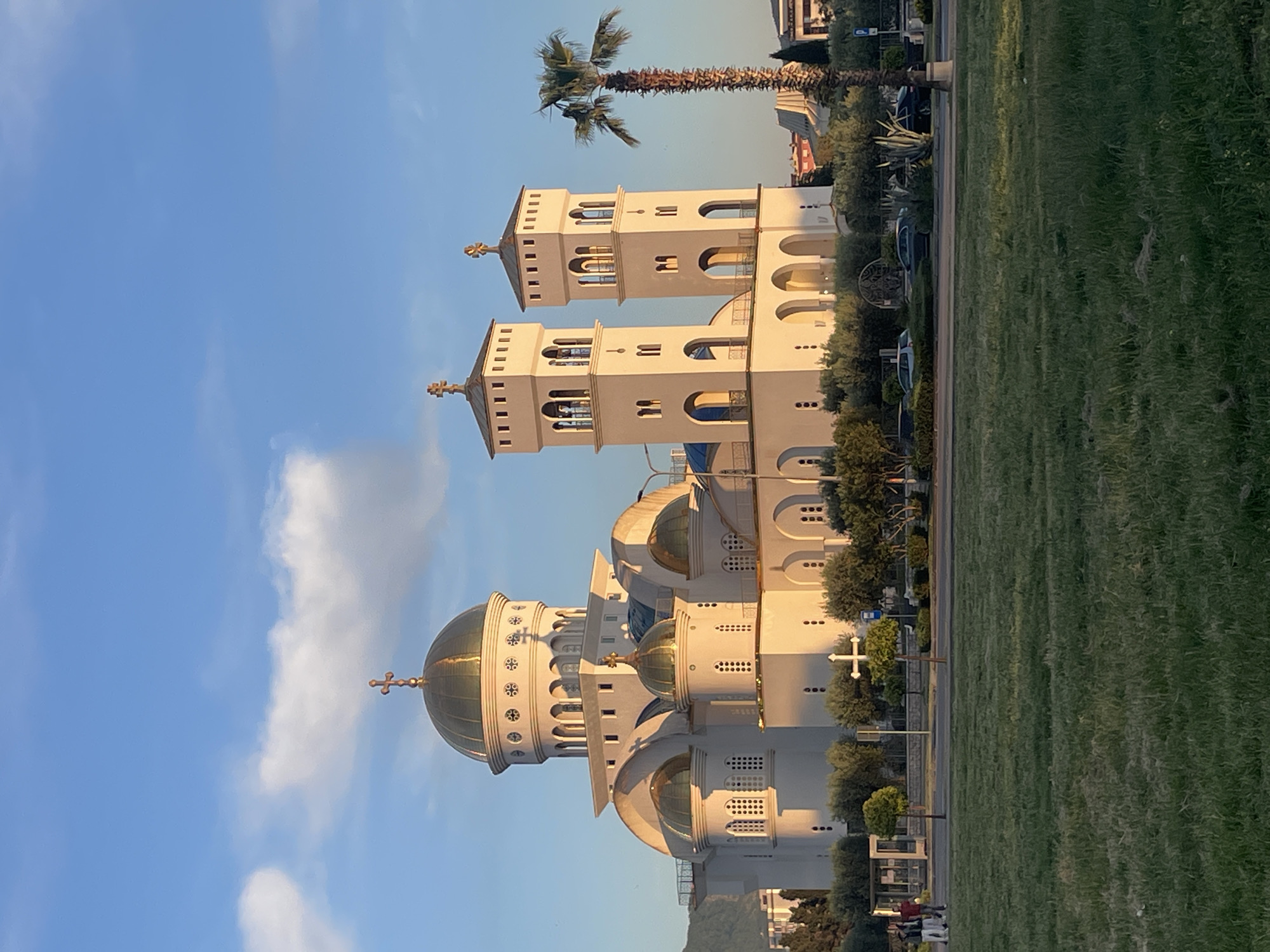 An image captures the majestic Cathedral of Saint Jovan Vladimir in Bar, Montenegro, under a clear, bright blue sky with a few scattered white clouds. The grand, white Eastern Orthodox cathedral, illuminated by the warm light of the golden hour, stands prominently on the left side of the frame. It features multiple golden-green domes, including a large central dome, and two prominent bell towers, all topped with golden crosses. The cathedral's architecture is characterized by intricate arches, windows, and decorative patterns. A smaller, white cross is visible on the church grounds to the right of the main building.
To the right of the cathedral, a wide, manicured lawn of green grass extends across the foreground and middle ground. A tall, slender palm tree rises behind the church's bell towers, adding a Mediterranean feel to the scene. In the background, beyond the grass and a faint road, other buildings and parked vehicles can be seen. The scene evokes a sense of peace and architectural grandeur. The strong sunlight and long shadows suggest either early morning or late afternoon. No clear human activity or discernible text is visible within the frame.