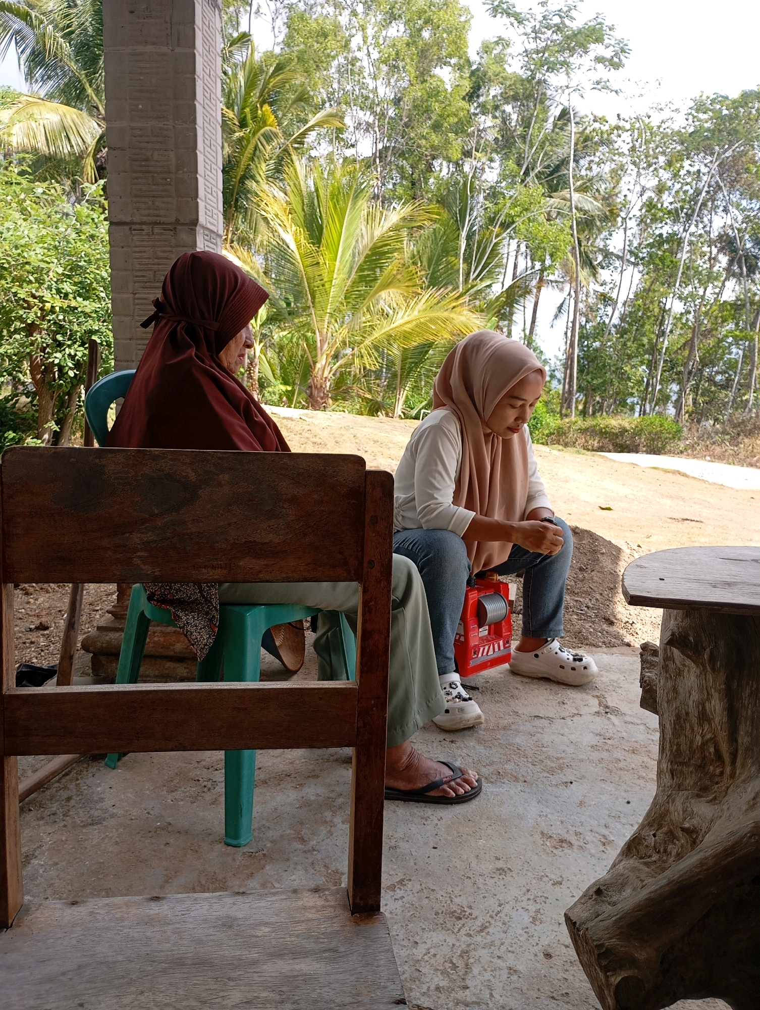 Two women are seated outdoors under a covered structure with a concrete floor. The woman on the left, wearing a maroon hijab and a printed skirt, is seated on a teal plastic chair. The woman on the right, wearing a tan hijab, white long-sleeved shirt, and blue jeans, is seated on a red plastic box. She is looking down at her hands. White clogs are on her feet. A wooden chair is in the foreground. Palm trees and other foliage are visible in the background. The location is the city of Ketapang, Indonesia.