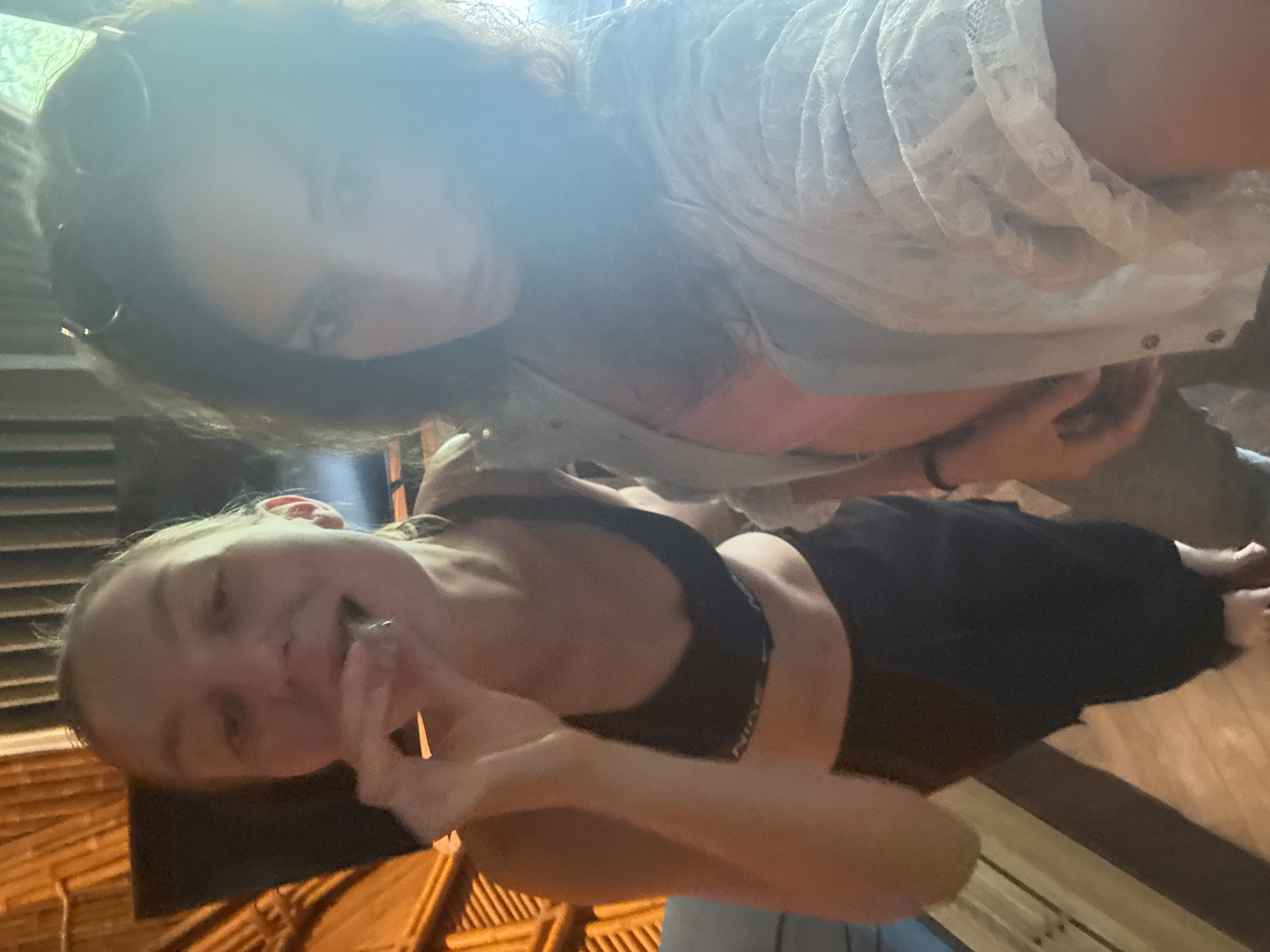The image depicts two adult females indoors. The specific location in the city of Mù Cang Chải, Vietnam, cannot be visually confirmed by the image content.
The woman on the left, positioned in the foreground, has light skin and light-colored hair pulled back. She wears a black sports bra, with a partial "Nike" logo visible on the band, and dark pants. She is barefoot. Her mouth is slightly open, and she holds a small, metallic, cylindrical object, resembling an e-cigarette or vaporizer, to her lips with her right hand. Her gaze is directed generally towards the camera.
The woman on the right, partially obscured by bright light and lens flare, has dark, curly hair with sunglasses perched on her head. She wears a light-colored, possibly pink, inner top beneath a white, long-sleeved, lace-patterned outer shirt. She holds a device, presumably a smartphone, to capture the image, looking directly at the camera.
The interior environment features a warm-toned, possibly bamboo or wood-paneled wall on the left. The floor consists of light wooden planks. A bright light source, likely a window or doorway, is present on the right, causing significant lens flare and obscuring parts of the background, though some blurred greenery is visible beyond the bright area.