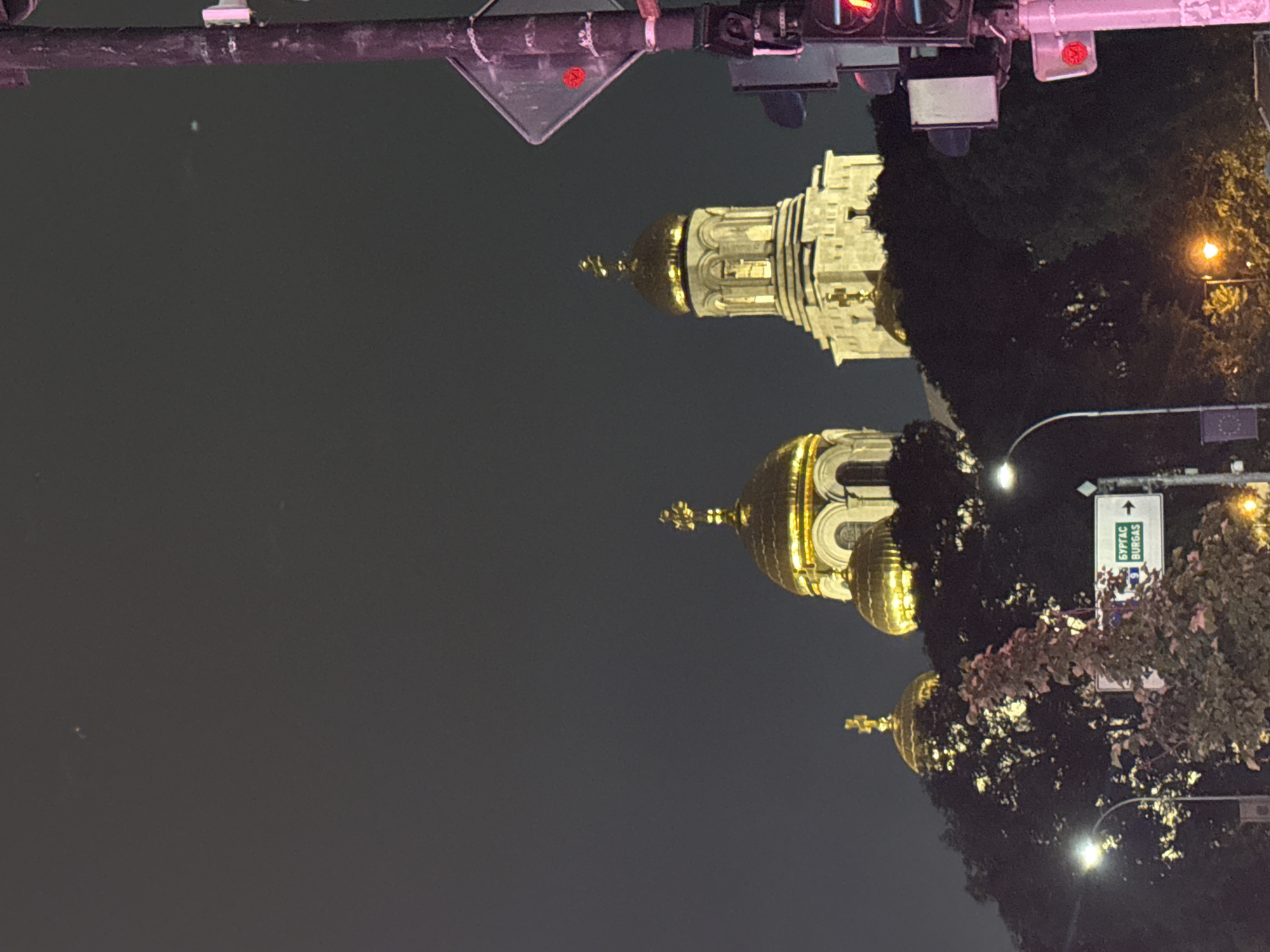 This detailed image captures an urban night scene dominated by three brightly illuminated golden-domed Orthodox cathedrals against a dark grey sky. The central cathedral is the most prominent, featuring a large golden dome topped with a cross, resting on a multi-tiered white stone drum with arched windows, which in turn sits on a substantial stone base. To the left, a smaller golden dome with a cross is partially visible behind dark foliage. To the right, another golden dome and cross are seen, belonging to a structure mostly obscured by a dark pole and trees.
The lower half of the image features dense, dark green trees and foliage, partially obscuring the lower sections of the cathedrals. Several artificial lights illuminate the scene: a bright white streetlamp is visible on the left, and an orange-toned streetlamp glows further right, closer to the foreground. In the midground, a white rectangular road sign is present, displaying text in both Cyrillic ("БУРГАС") and Latin script ("BURGASS"), along with the number "9" and an arrow pointing to the right. On the far right, a dark, thick utility pole stands vertically, holding a set of red traffic lights, indicating a stop. Above the traffic lights on the pole, a white rectangular sign with a red border and a small red circular emblem is mounted. The sky above is uniformly dark grey, with a few minute, faint light specks discernible.
The location is identified as Varna, Varna Province, Bulgaria, based on the distinctive architecture resembling the Dormition of the Theotokos Cathedral and the road sign indicating "Burgas," a major city to the south along the Bulgarian Black Sea coast.
