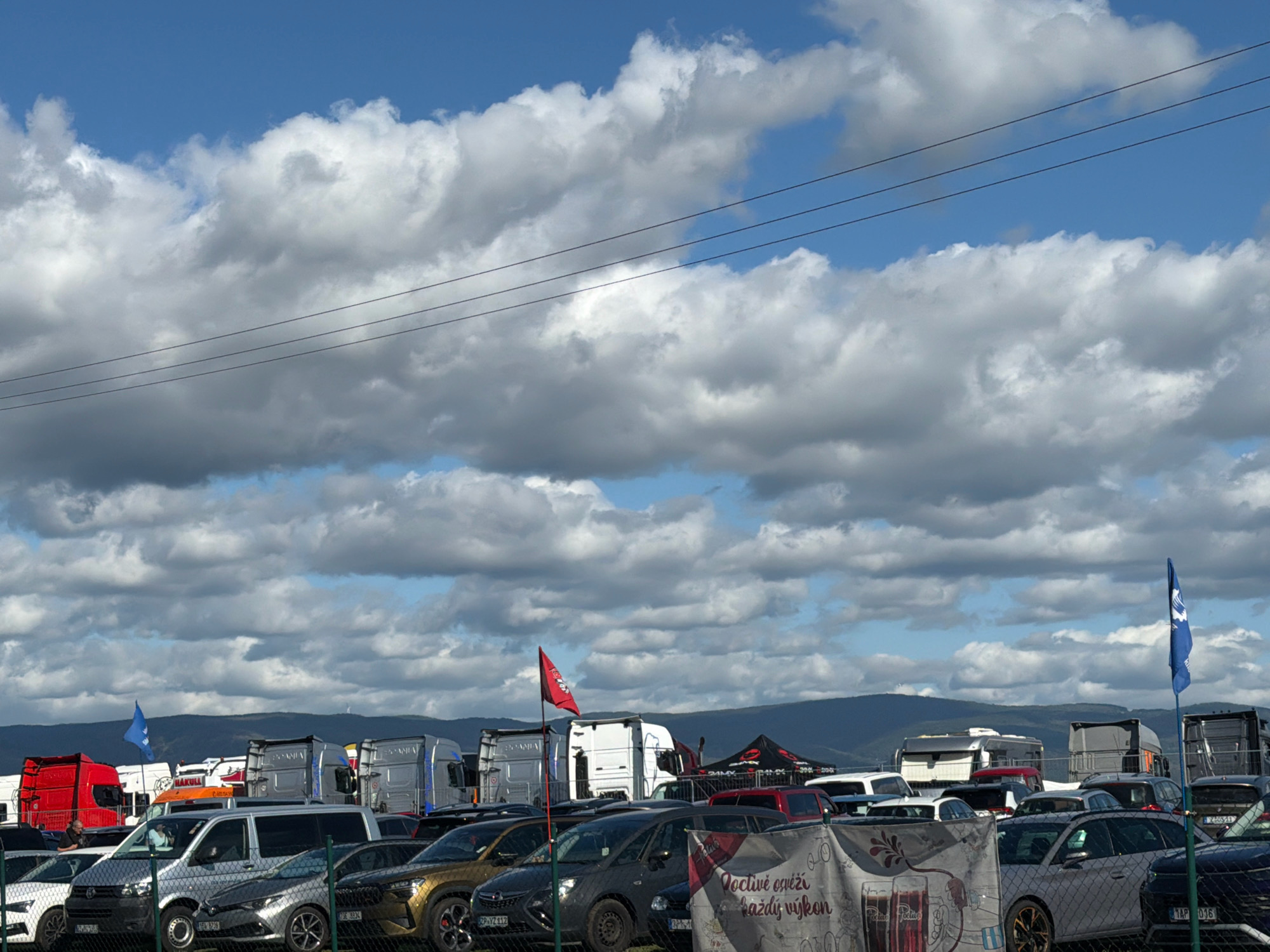The image shows a large outdoor parking lot filled with various vehicles, including trucks, vans, and cars, under a cloudy sky. The scene appears to be at a venue or event, possibly related to motorsports, given the presence of multiple large trucks and a red flag with a crest that might signify an organization or team. In the foreground, a fence separates the viewer from the parking area. There are several trucks, including red, white, and silver ones, parked in rows. Cars are also visible, parked behind the trucks. In the background, hills or mountains are discernible. The sky is a mix of blue and white clouds, suggesting a partly sunny day. The overall impression is of a busy, outdoor gathering or event. The location of Most, Czechia is not visually confirmed by any signage in the image.