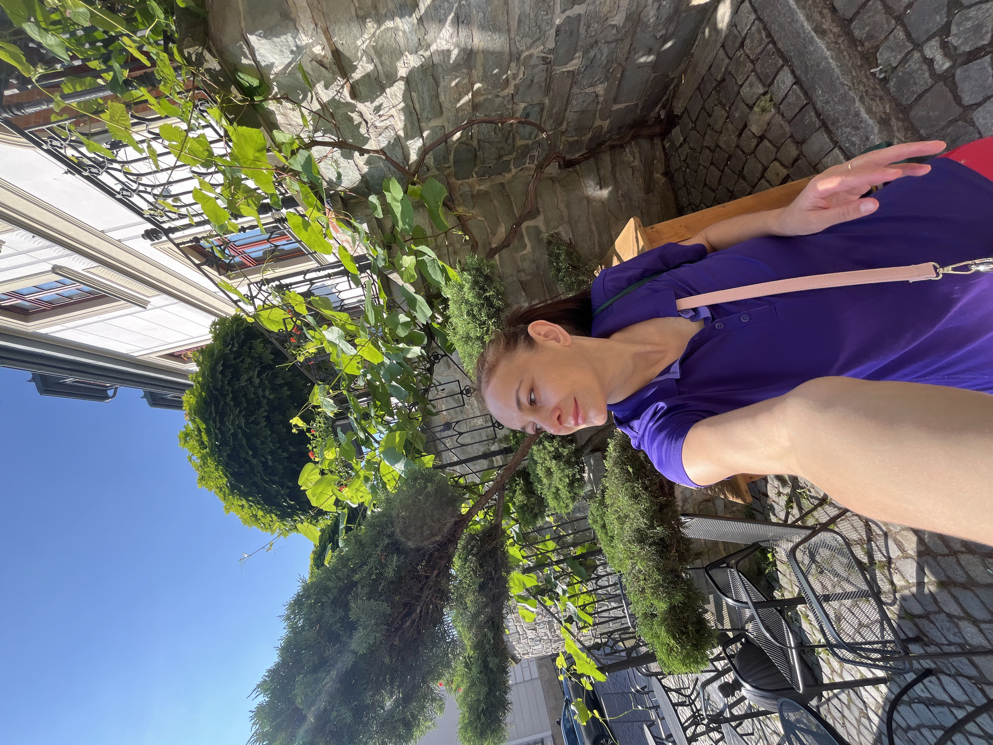 A woman is sitting on a wooden bench outdoors, with a vine and greenery growing on a stone wall behind her. She is wearing a purple polo shirt and a light pink strap is visible across her shoulder. She has a ring on her left ring finger. The ground is paved with cobblestones, and a metal table and chairs are visible in the foreground. The scene appears to be a sunny day, likely in the afternoon, given the shadows cast by the foliage and furniture. The woman is looking to her right, with a slight smile on her face, suggesting a relaxed or content mood. The backdrop includes a building with windows and architectural details, hinting at an urban or historical setting. There is no visible text in the image.