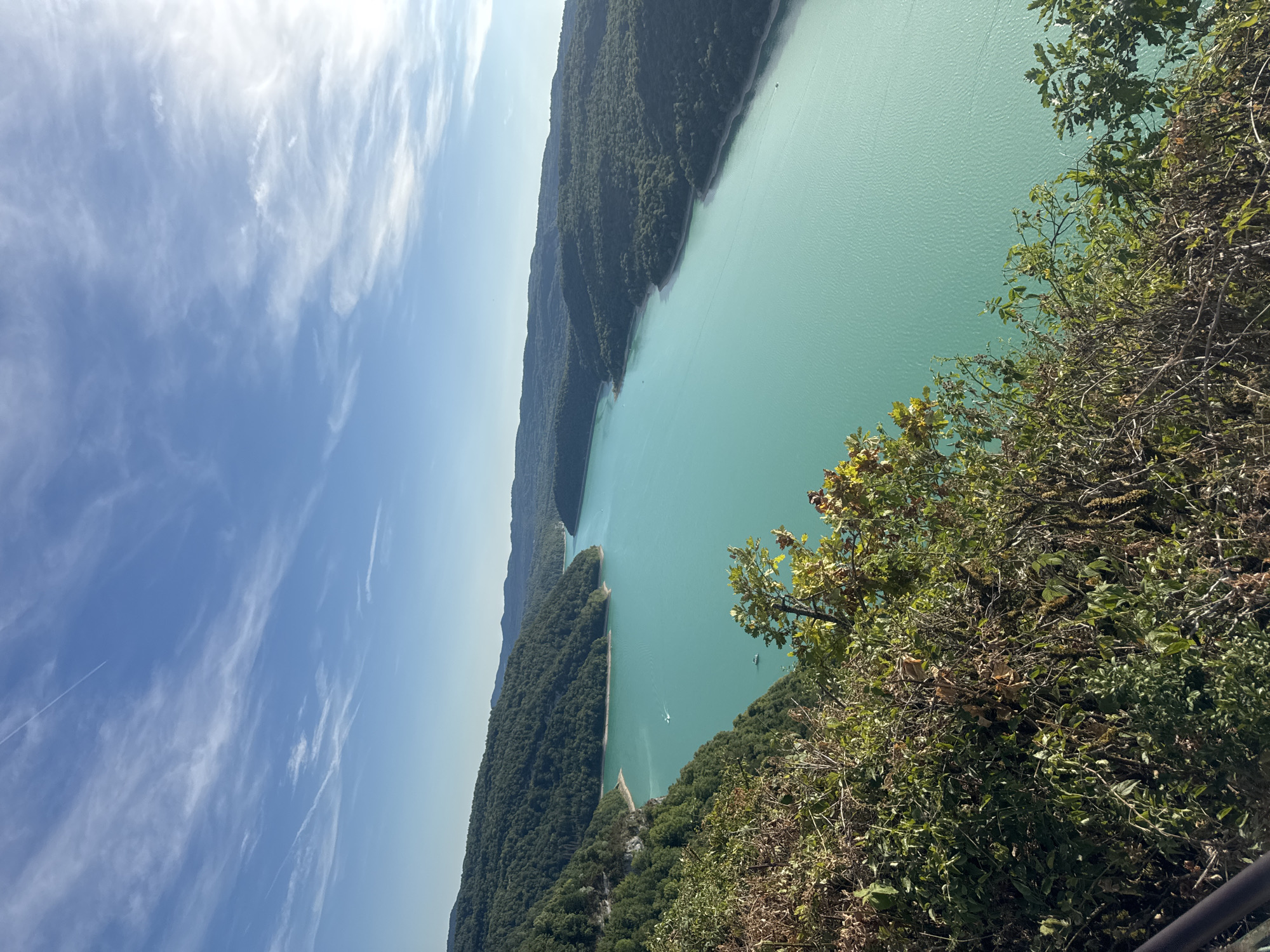 The image captures a serene panoramic view of the Lac de Vouglans, a man-made lake located in the Jura region of France, near the commune of Moirans-en-Montagne. The viewpoint is from a slightly elevated position, overlooking the vast expanse of turquoise water. Lush, green, tree-covered hills flank the lake, their slopes gently receding into the distance. The sky above is a clear, vibrant blue, dotted with wispy white clouds and traversed by faint contrails from aircraft. In the foreground, the edge of dense green foliage and dry branches creates a natural frame for the scene. A few small boats are visible on the tranquil water, leaving subtle wakes. The overall impression is one of natural beauty and peacefulness.
