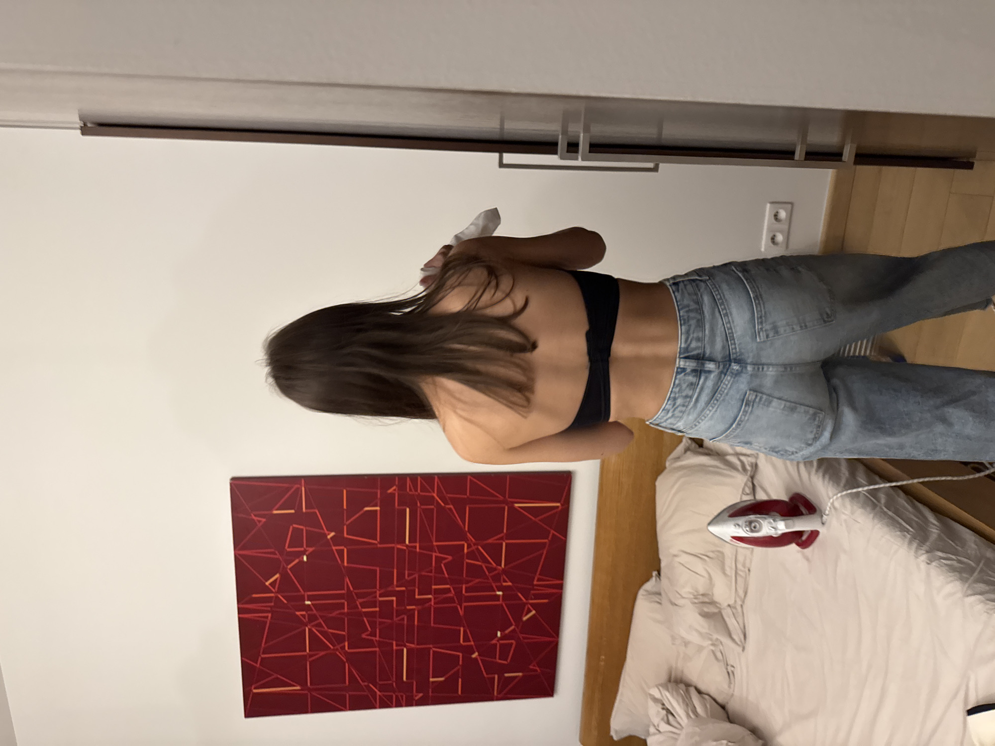 A woman in a black bra and jeans is ironing clothes on a bed in a room with a painting on the wall. The painting is a red abstract design with yellow accents. A red and white iron is on the bed next to her. The location is Unknown, Unknown.