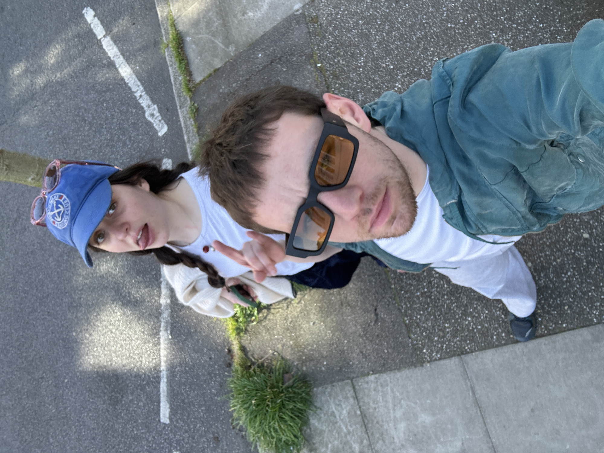 Two individuals, a male and a female, are present in a high-angle selfie photograph. The male, wearing sunglasses and a blue jacket over a white shirt, holds the camera. The female, positioned to the left of the male, wears a blue cap with pink sunglasses placed on top, a white shirt, and has her hair in a braid. She is holding up a peace sign with her fingers.
The background includes a street with white painted lines and patches of grass growing along the curb. The location appears to be an urban environment, most likely in London, United Kingdom due to the style of dress and the common urban landscape features. The photo seems to be taken at a street level.