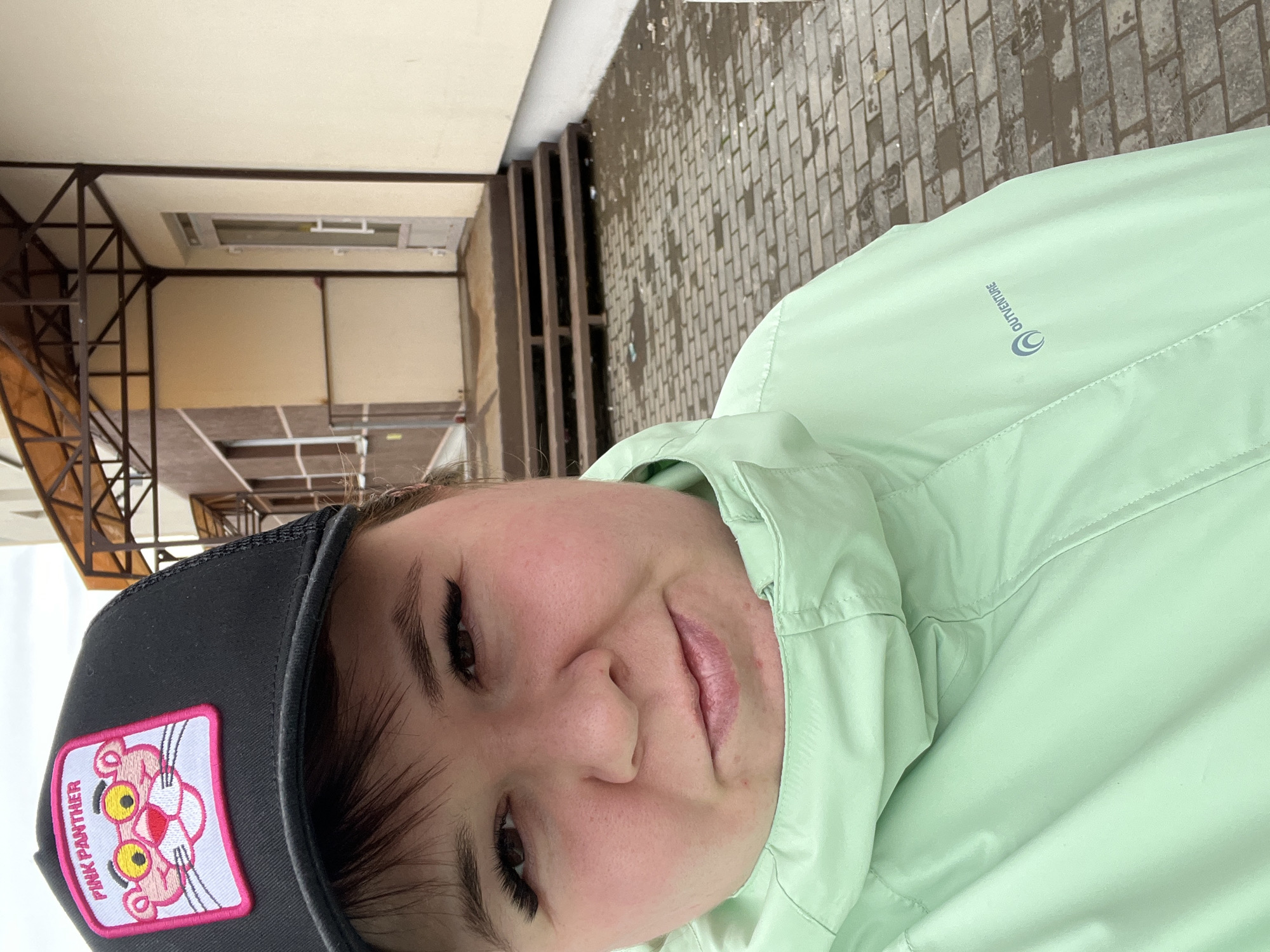 A person wearing a black baseball cap with a "Pink Panther" patch and a light green Outventure jacket is posing for a selfie. The background includes a building with brown and beige exterior walls, visible through a metal structure. A stone-paved walkway is also partially visible. Based on the information provided, the location is Chaikovsky, Russia.