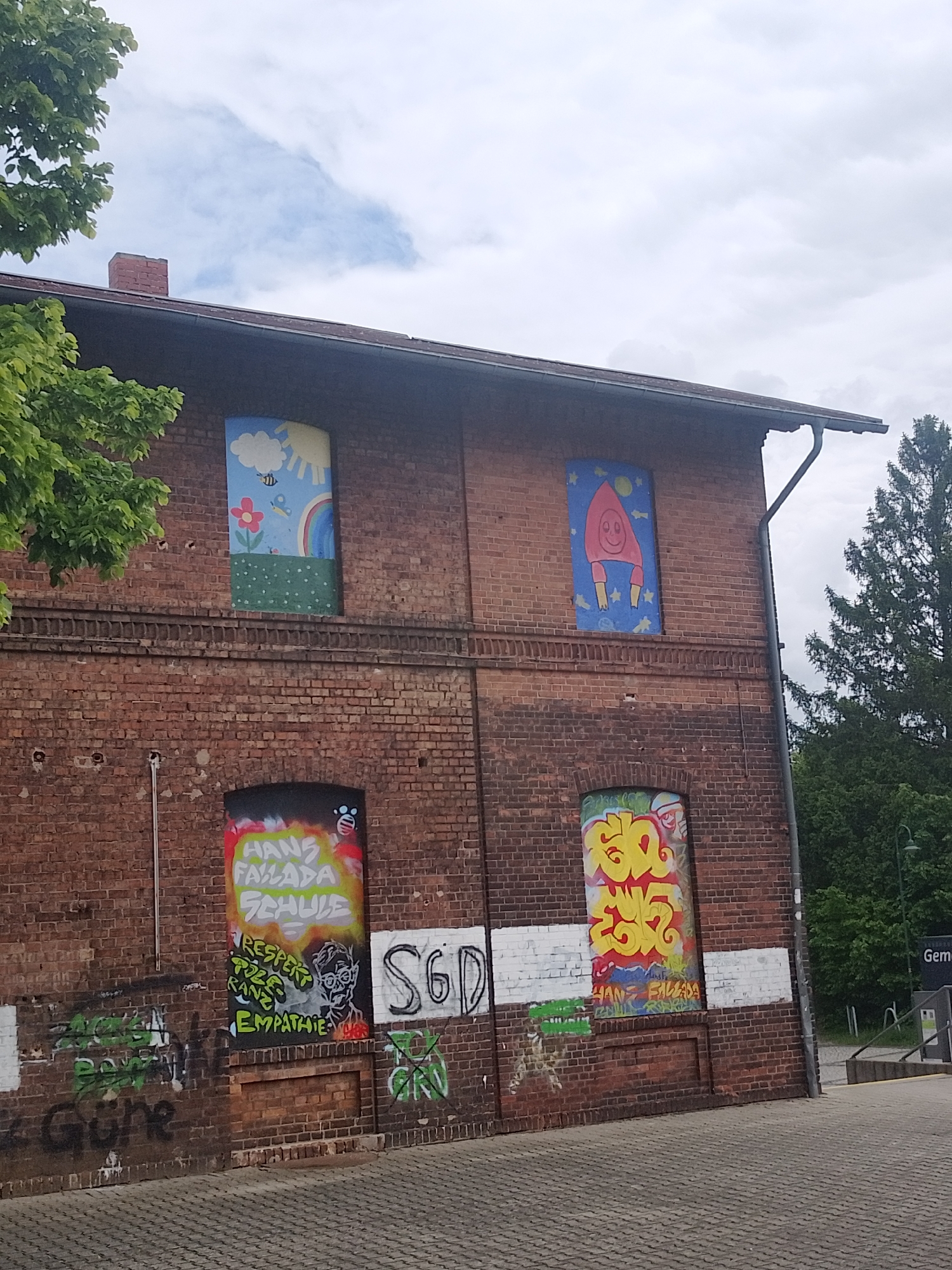 The image shows a two-story brick building with graffiti and murals covering the windows and lower walls. The upper windows are painted with childlike images: one with a sun, clouds, flowers, a rainbow, a bee, and a butterfly; the other with a red rocket ship against a blue background with stars and a moon. The lower windows and walls have extensive graffiti in various colors, including green, yellow, red, white, and black. Some identifiable words within the graffiti include "HANS FALLADA SCHULE," "RESPEKT," "TOLERANZ," and "EMPATHIE." A small area of white paint is visible along the base of the building. The building is surrounded by pavement and trees. A sign with the word "Geme" is visible in the background to the right of the building. The location is in the city of Unknown, Unknown.
