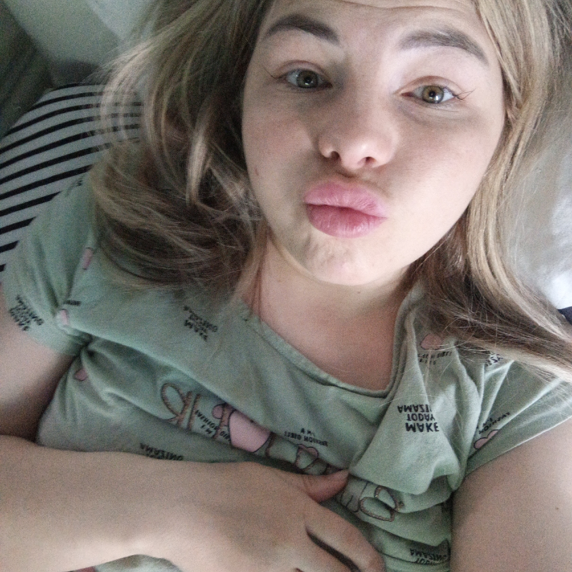 A fair-skinned woman with blonde hair lies on a striped pillow, wearing a light green pajama top with pink heart details and text. She is puckering her lips towards the camera. The setting is unknown.