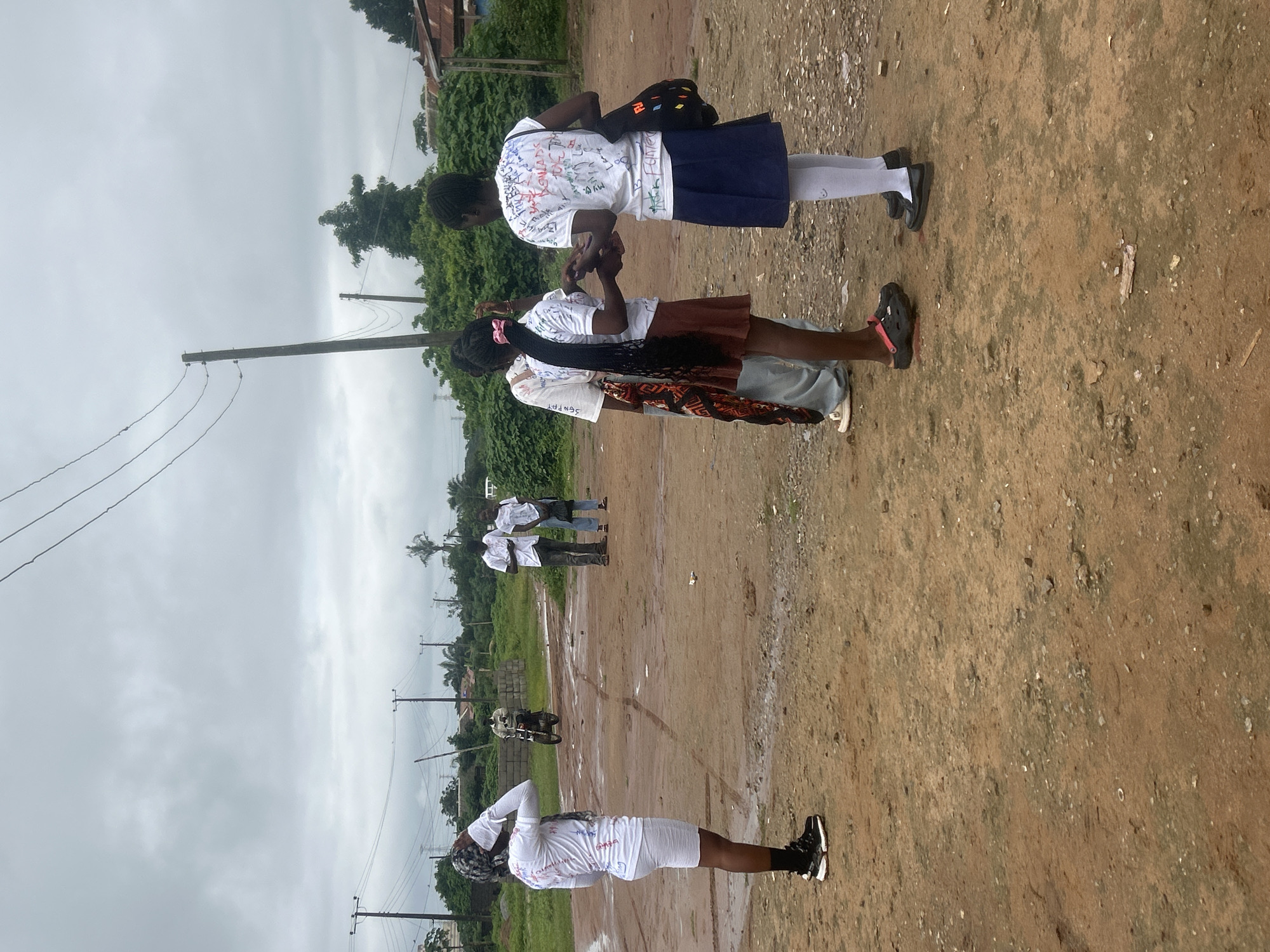The image depicts a group of young people, likely students, on what appears to be a dirt field in Ikorodu, Nigeria. The sky is overcast, suggesting a cloudy day. In the foreground, two individuals in white shirts and skirts are interacting. The person on the left has a long braided hairstyle with a pink ribbon. The person on the right is wearing a dark blue skirt and has a backpack slung over their shoulder. Both are wearing white shirts that are decorated with various writings, possibly messages or signatures, typical of graduation or farewell attire.
Further back, other individuals in white shirts are visible, some standing in pairs. One person in the mid-ground is wearing a white shirt and white shorts, with their hands behind their head. Another individual in the background is leaning down, and a motorcycle is parked nearby. The setting is rural or semi-rural, with lush green vegetation and trees visible in the distance, as well as power lines and poles. The ground is predominantly brown dirt, with some patches appearing wet, indicating recent rainfall. The overall atmosphere seems casual, possibly marking the end of a school term or a significant event for the students.