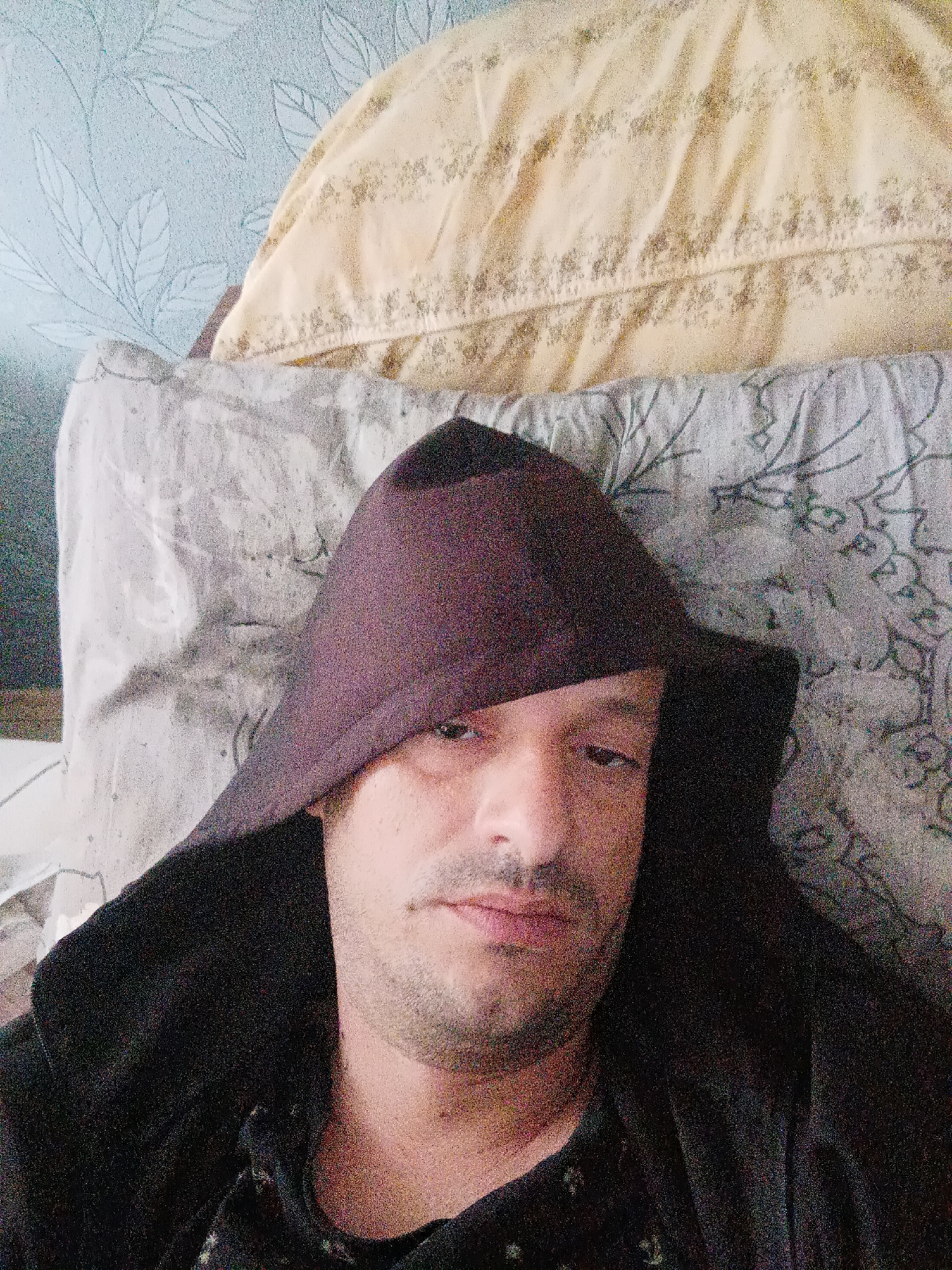 A close-up, medium-shot photograph depicts a man with light skin and stubble lying on a pillow or bed, possibly indoors. He is wearing a dark-colored robe or hooded garment. The background includes patterned bedding and a wall or headboard with a light blue floral pattern. There is no discernible information to place the scene in Burgas, Bulgaria, or any city for that matter. The primary subject is the man. The setting seems to be a bedroom based on the visible bedding. The action is the man lying down. There are no clear interactions.