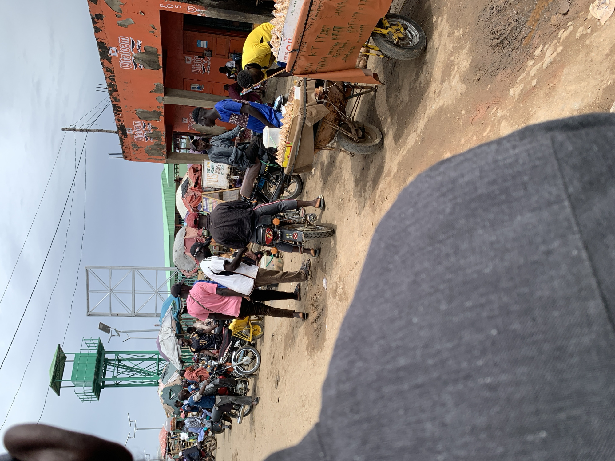 The image depicts a busy street scene in Keffi, Nigeria. A line of people on motorcycles are waiting along the roadside next to buildings with signs that include the brand "Vitafoam." Some of the motorcycles are positioned near a makeshift market stall selling food. There is a water tower visible in the distance. The image is taken from a close, obstructed viewpoint.