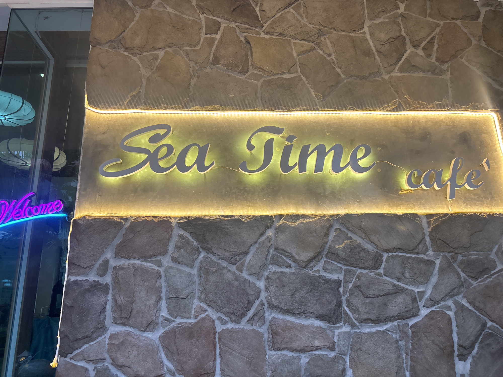 In the city of Chalong, Thailand, a brightly lit sign reading "Sea Time cafe" is mounted on a stone wall. The sign is illuminated with a warm yellow light, and the letters are a sleek, modern design. To the left of the sign, a neon pink "Welcome" sign glows in front of a glass storefront, reflecting the lights of the interior. The stone wall is constructed from irregular, rough-hewn stones, with light-colored grout filling the spaces between them. A strip of small, white LED lights runs along the top edge of the sign, casting a soft glow upwards onto the stone. The overall impression is of a welcoming and stylish establishment.