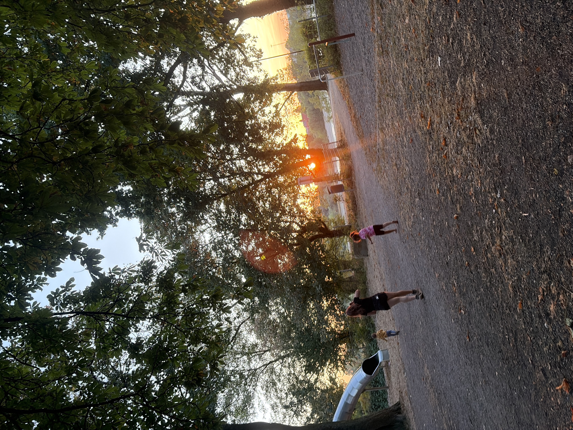 The image depicts a park scene at sunset, with a prominent metal slide on the left. Three individuals are visible: a woman in a black top and shorts, a young child in a yellow shirt, and another child in a pink top. They appear to be in motion, possibly playing or running on the gravel ground. The background features mature trees with dense foliage, and the setting sun casts a warm glow, creating lens flares. The overall environment suggests an outdoor recreational area in an urban setting. This image was taken in the Sportpark Duisburg, Germany.