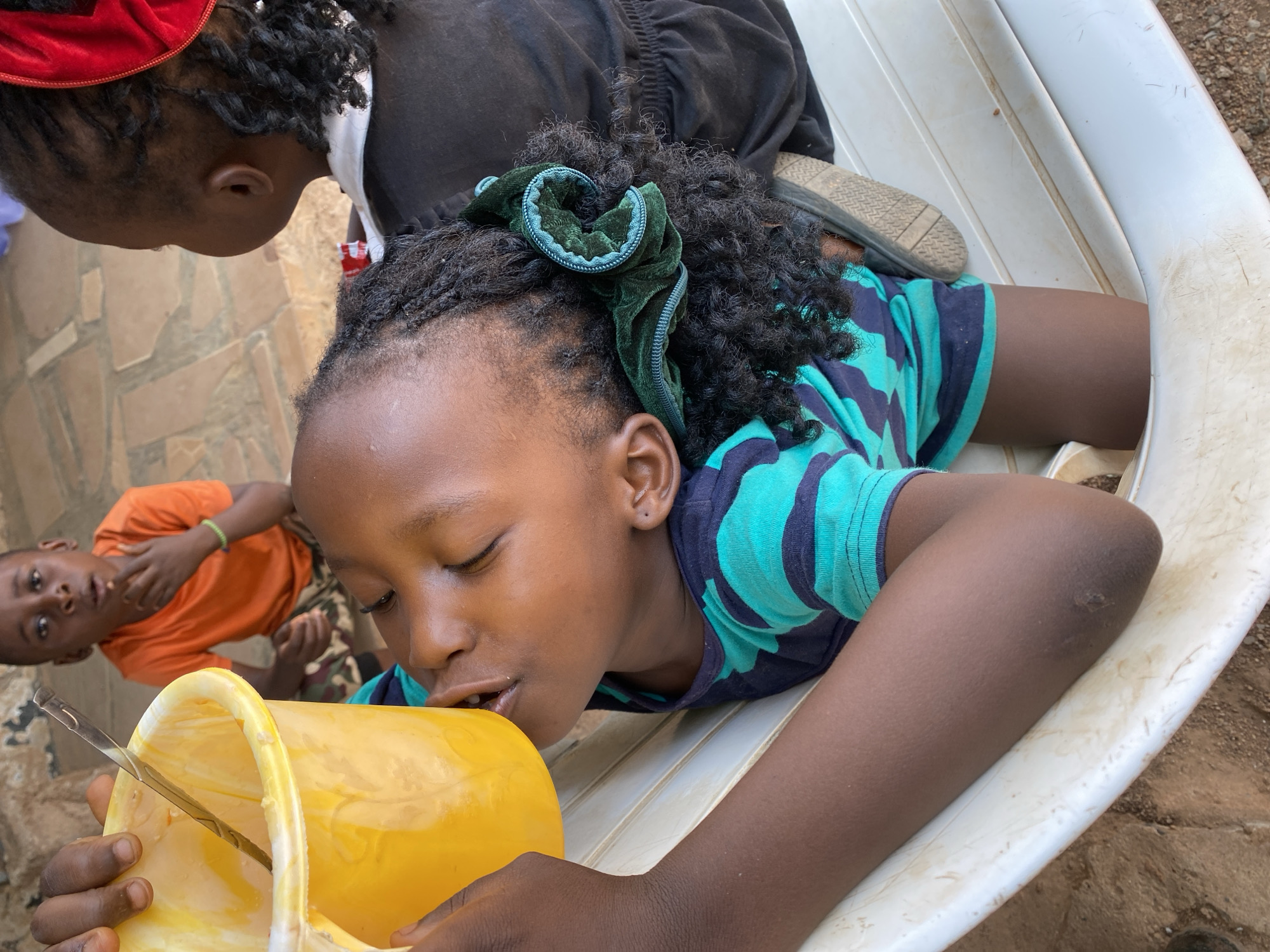 A young Black child is seated in a white plastic chair, tilting a yellow plastic bucket to their lips, apparently drinking its contents with the aid of a metal spoon. The child has braided hair with a green scrunchie. They are wearing a striped teal and navy shirt. Another child wearing an orange shirt can be seen in the background. A third child with a red hairband is seen from above. The location is likely Kuru Karama, Nigeria.
